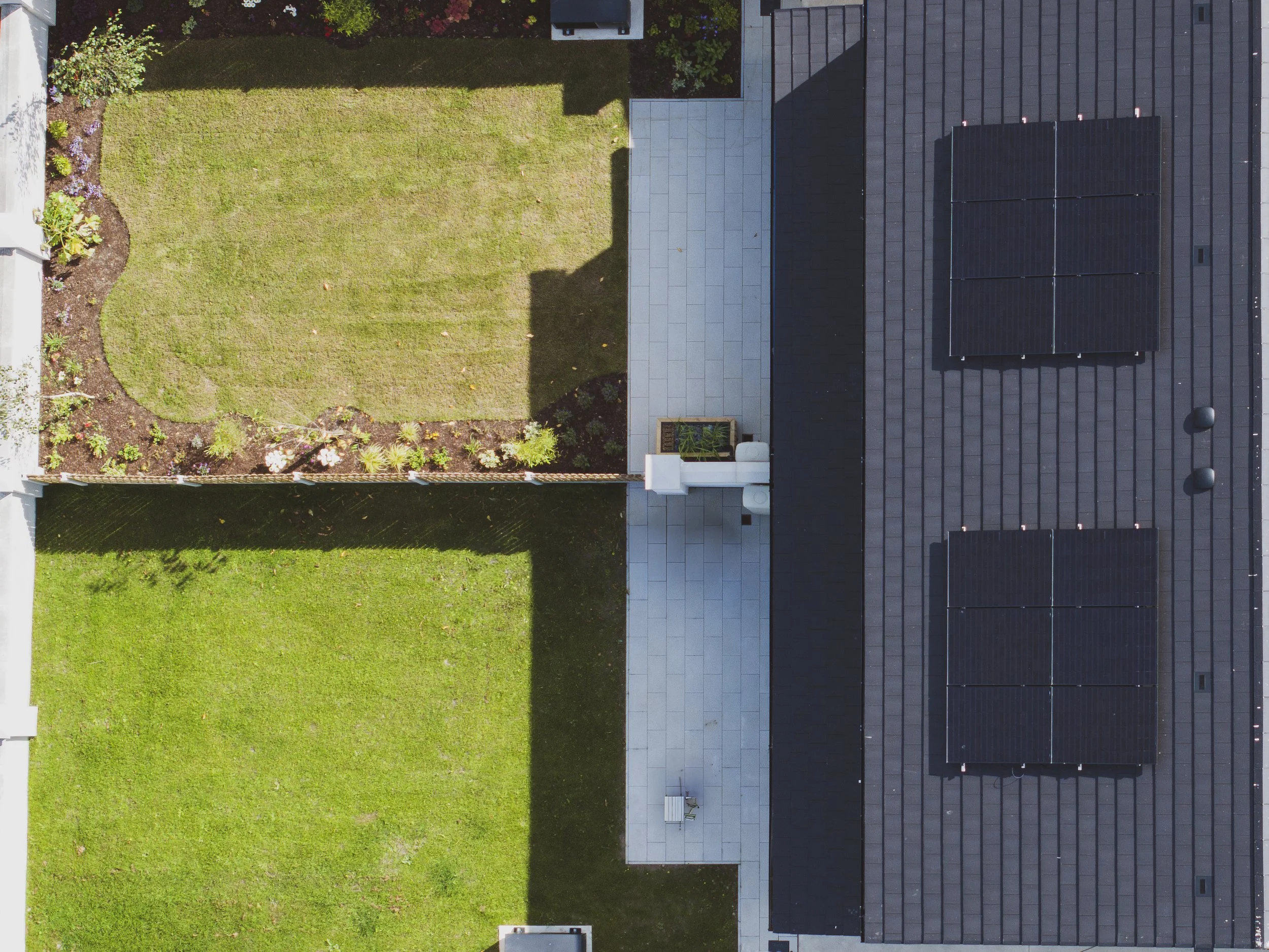 An aerial view of a backyard with a garden on the left and a patio with solar panels on the right. The garden has a grassy area with some plants and flowers along the border. The patio has a tiled floor, a small garden bed, and some vents or skylight
