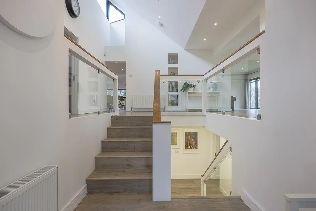Interior view of a modern, multi-level home with wooden stairs, glass railings, and white walls, featuring artwork and decorative items.