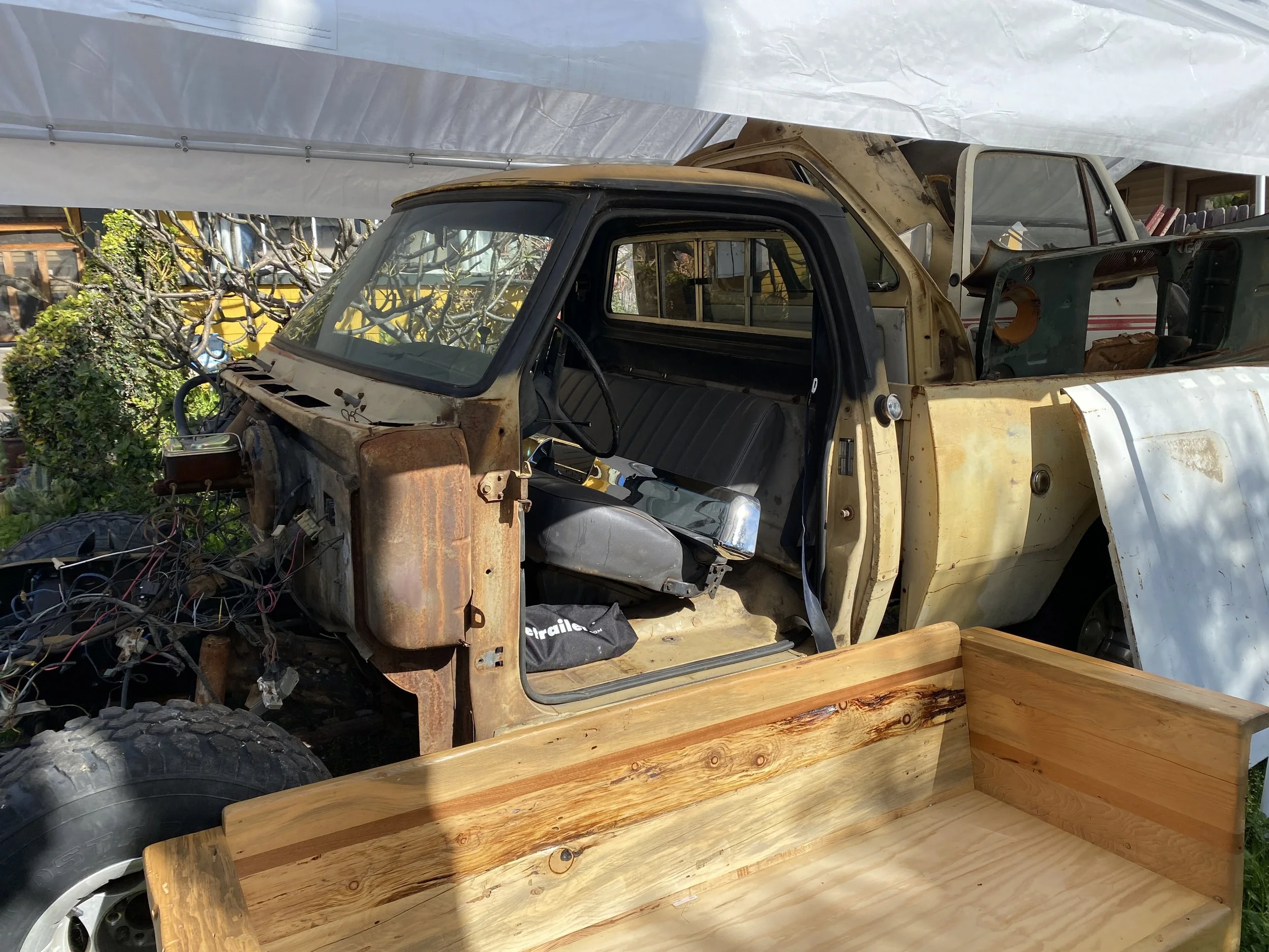 A vintage partly disassembled truck with a yellow body and black interior, parked outdoors next to green bushes and trees, with a white canopy partially covering it, and a wooden bench with a natural finish in the foreground.