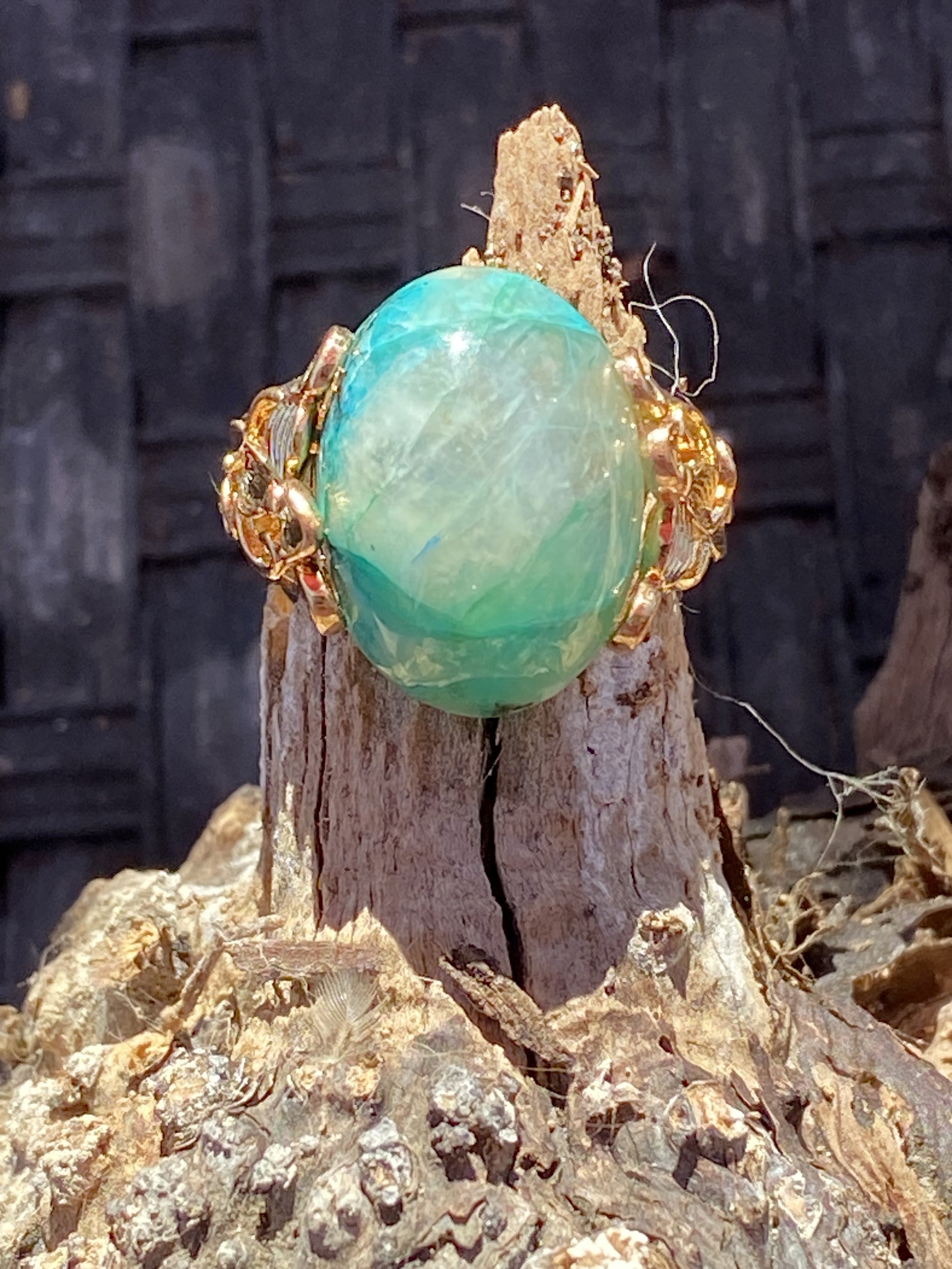 Natural amazonite gemstone ring with a vibrant blue-green cabochon set in an ornate gold-tone setting displayed on weathered wood.