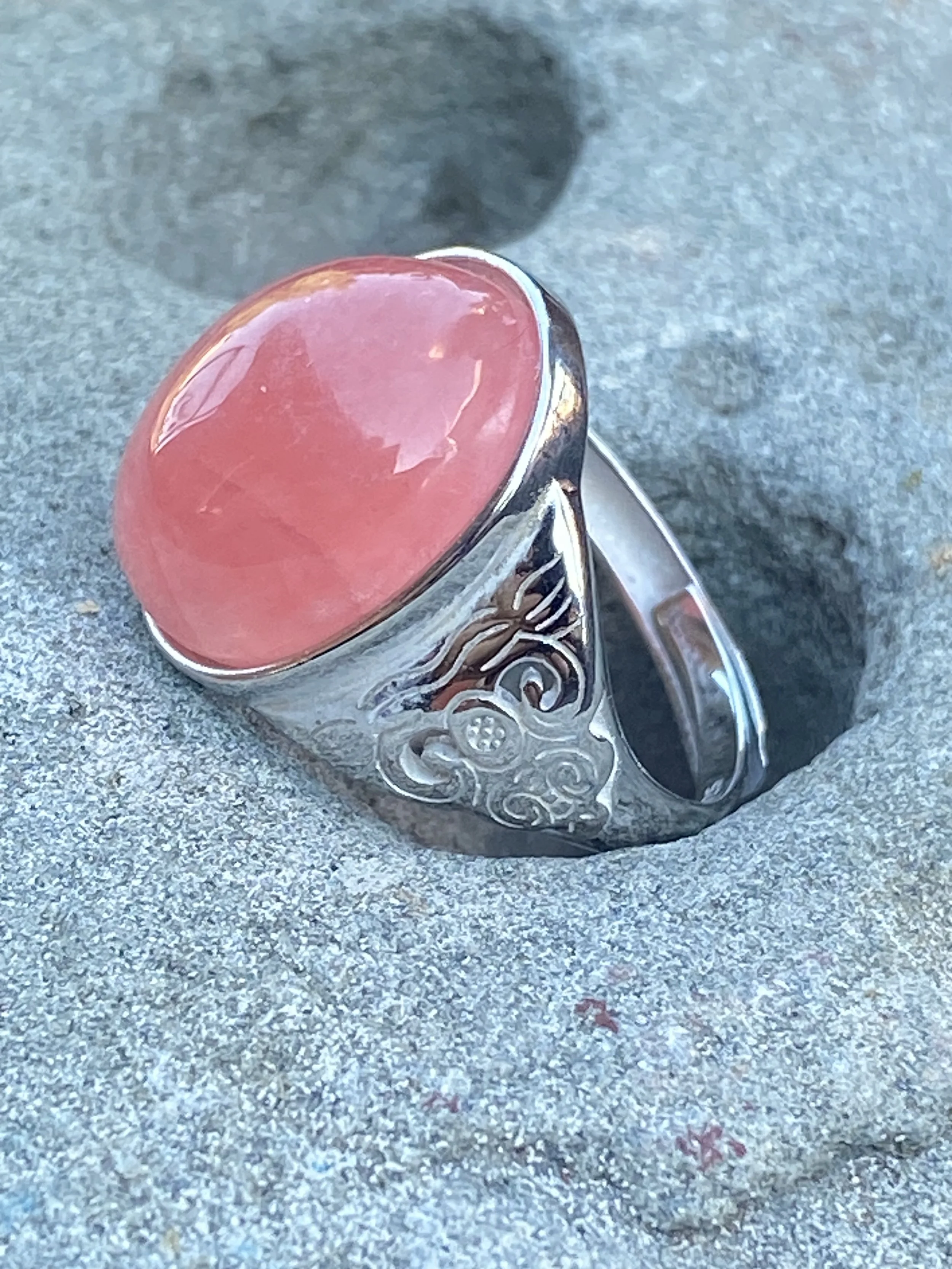 Pink cabochon gemstone ring set in an engraved sterling silver band displayed on natural stone.