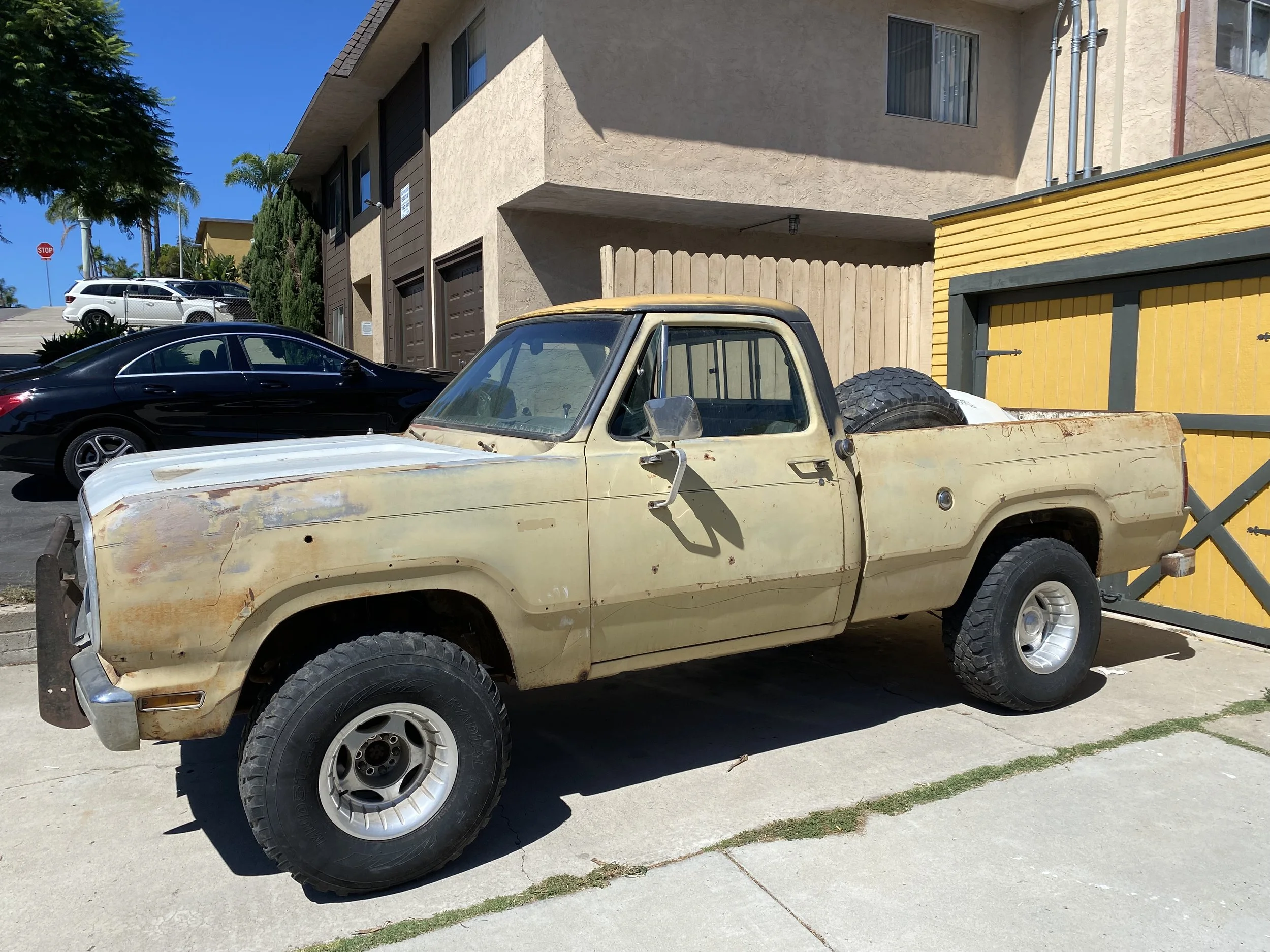 An old, dusty beige pickup truck parked in front of a residential building with yellow and grey garage doors, next to a black car, and other vehicles parked further up the driveway, with trees and a stop sign in the background.