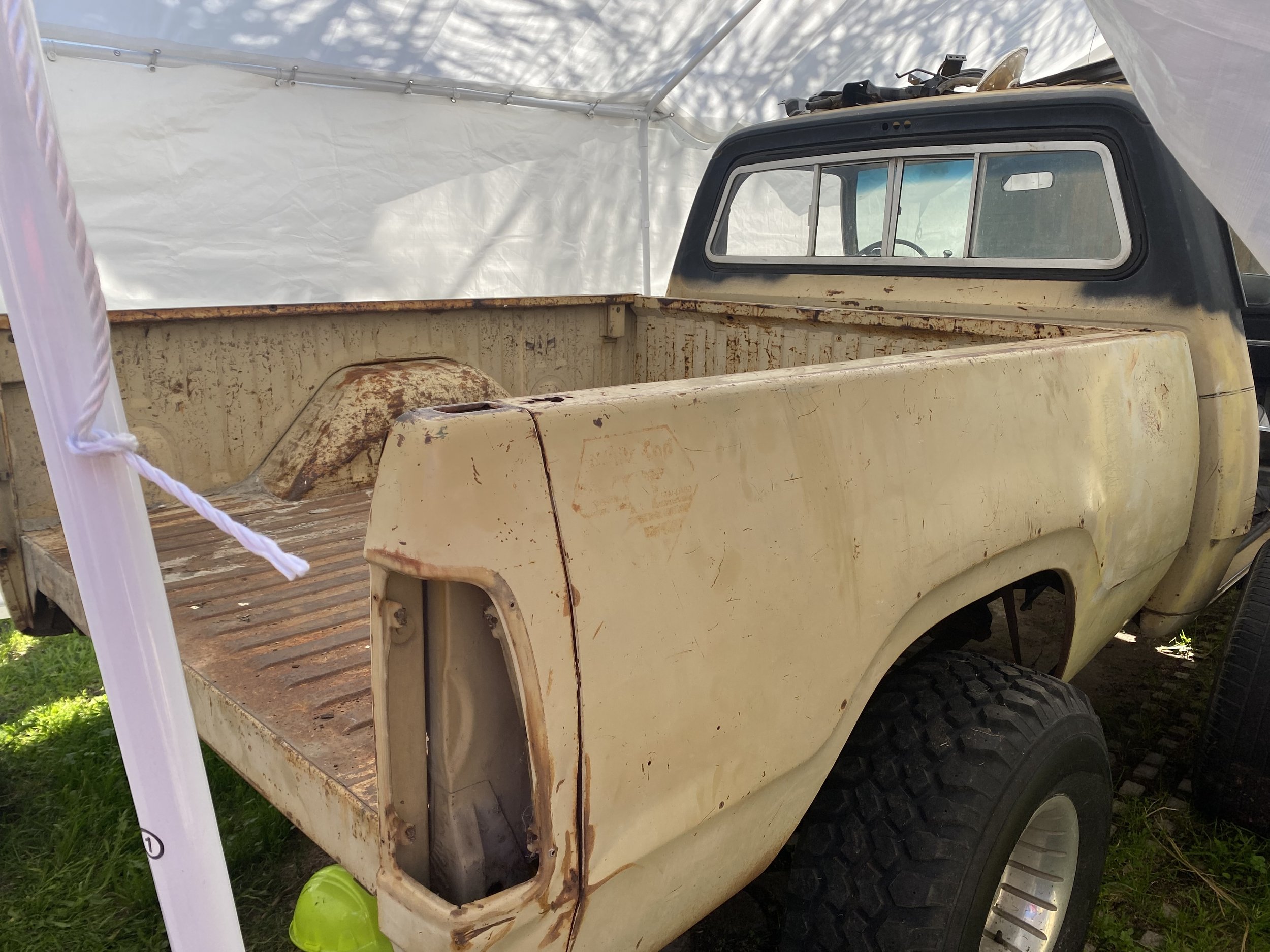An old, rusty pickup truck with a dirty, beige body and black roof in a tent or covered area, with some tools or equipment on top, and a green ball on the grass nearby.