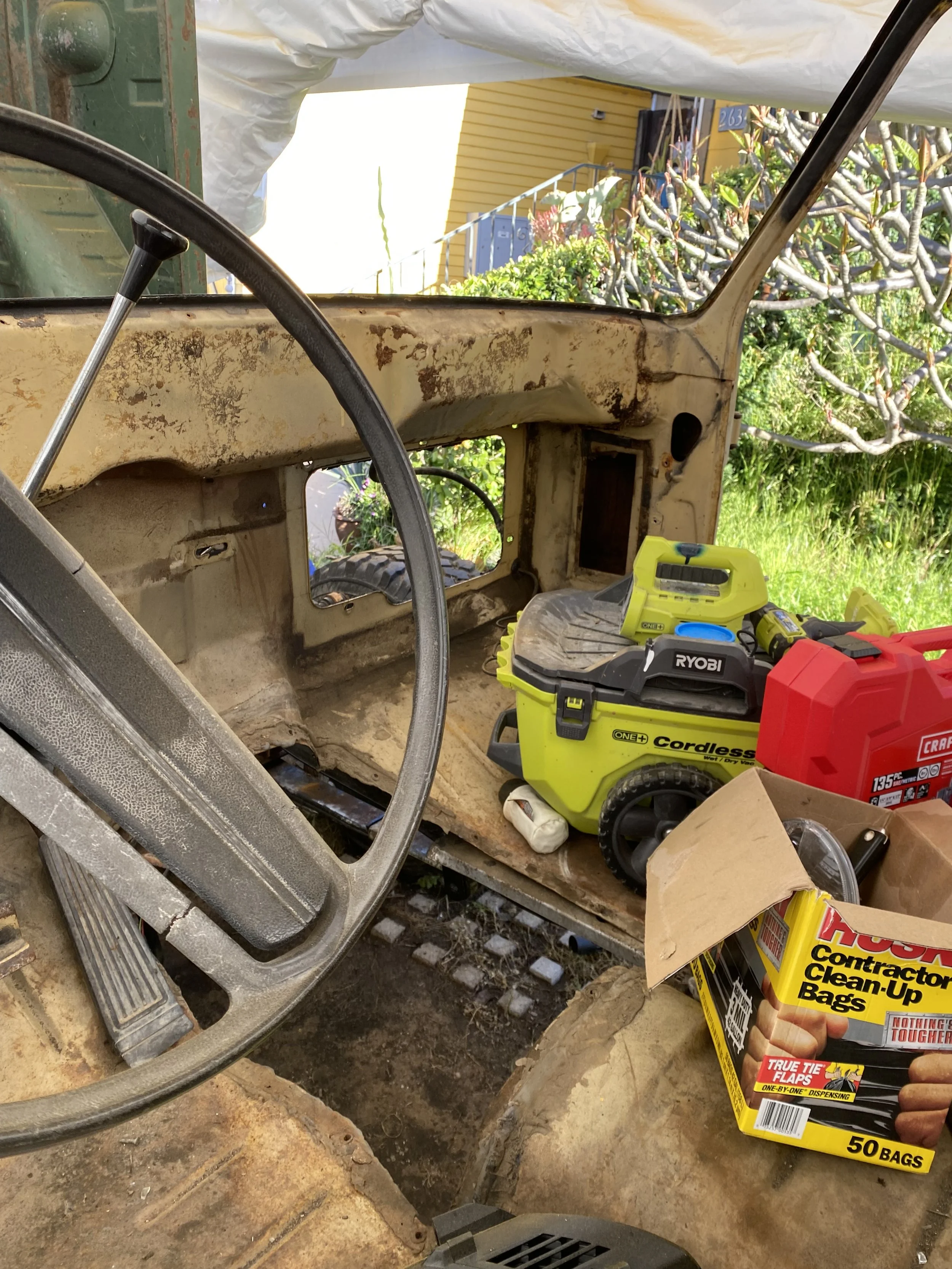 View from inside a rusted and partially disassembled vintage vehicle, with tools and cleaning supplies around, and the outdoors with greenery and a yellow house visible through the window.
