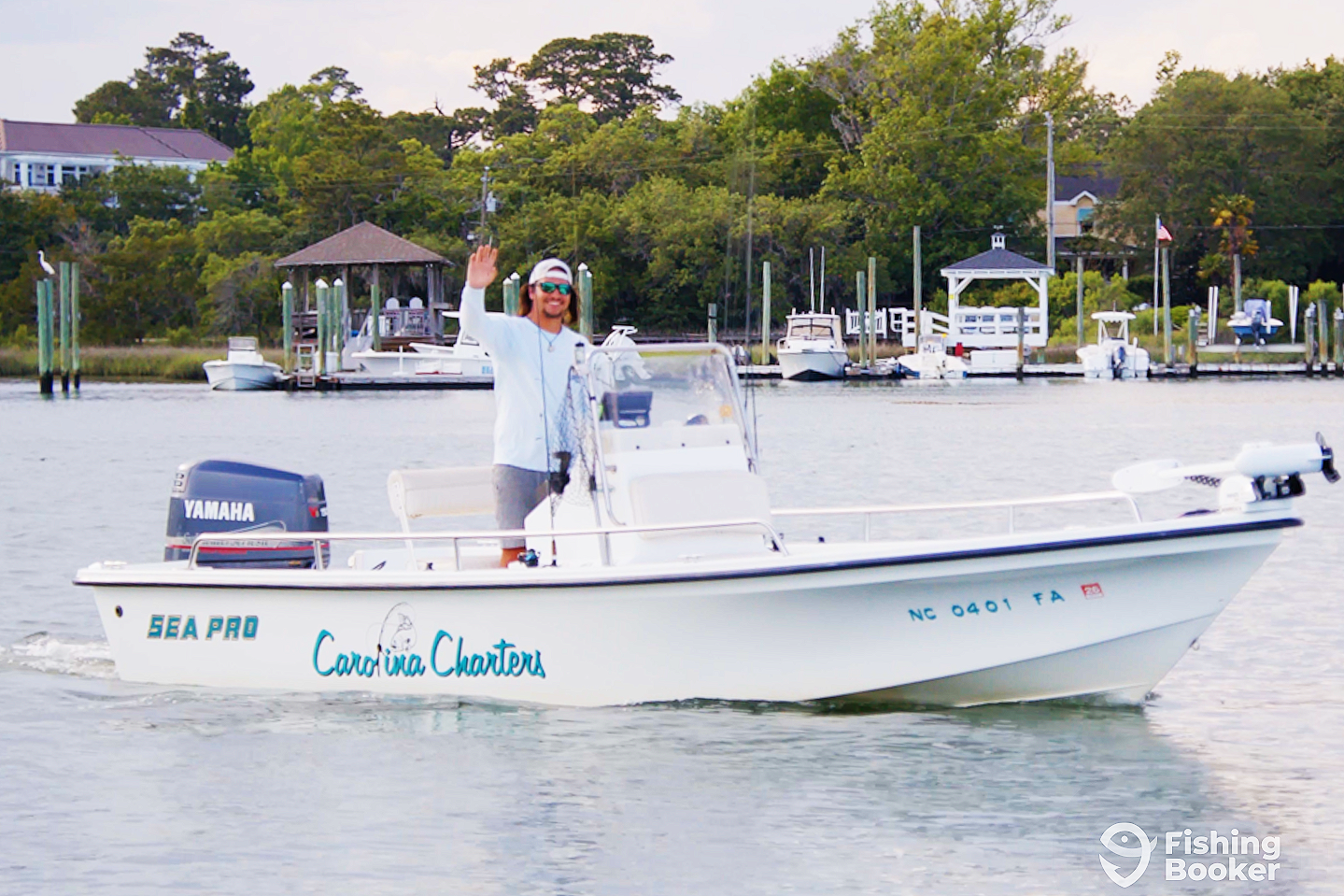 A man waving from a small white fishing boat on the water with trees and houses along the shoreline in the background.