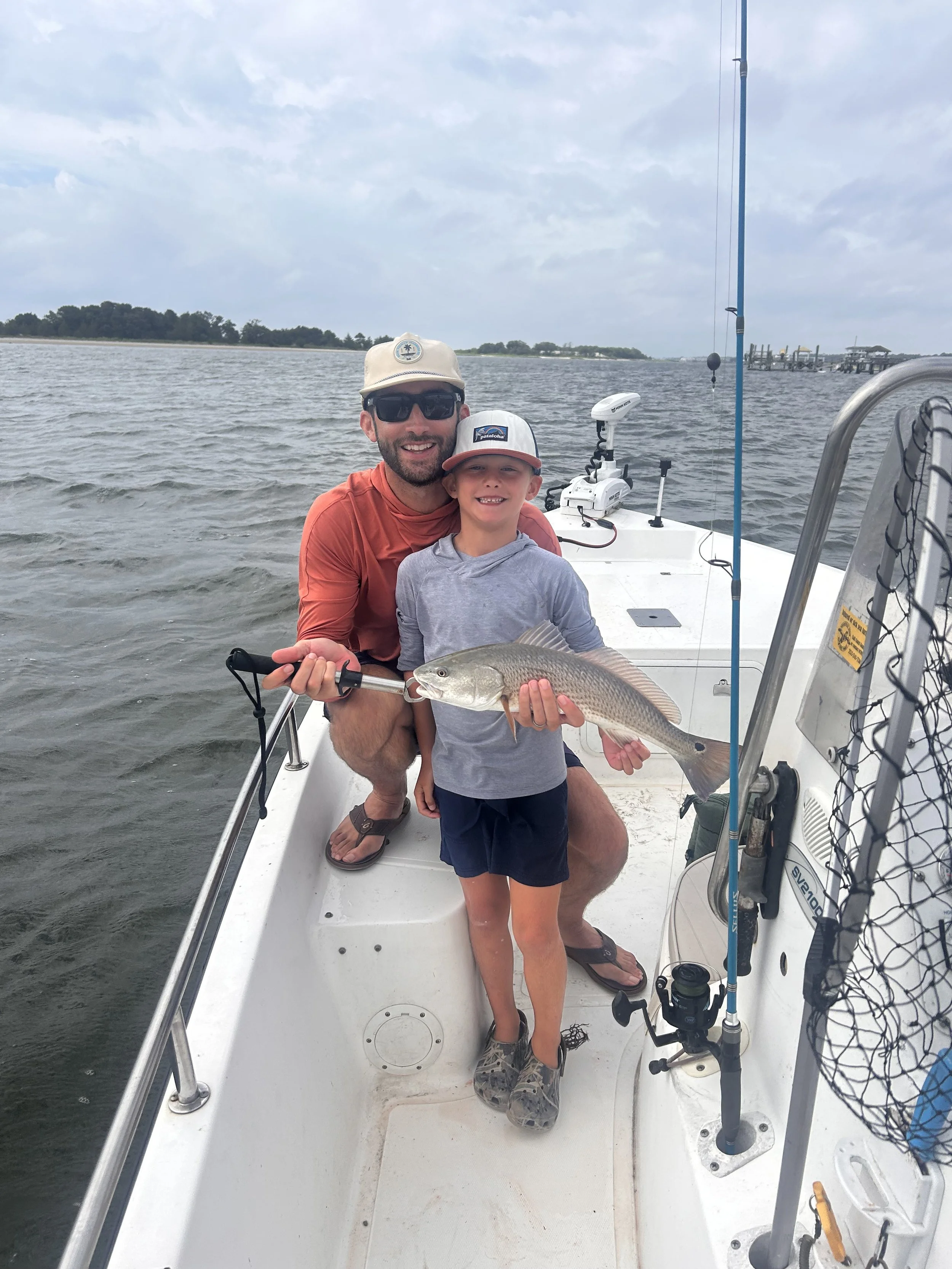 A man and a boy on a boat holding a fish they caught, smiling at the camera with water and a cloudy sky in the background.