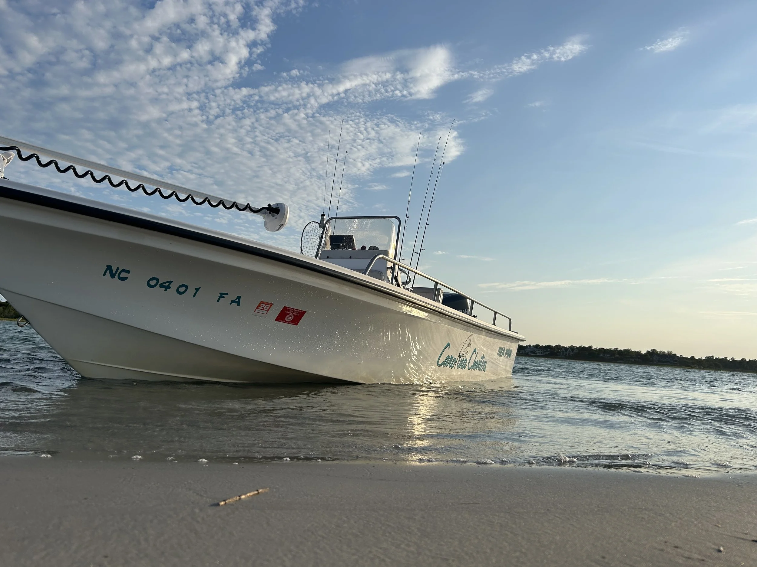 A white boat resting on the shore near the water with a few fishing rods. The sky is partly cloudy, and the water is calm.