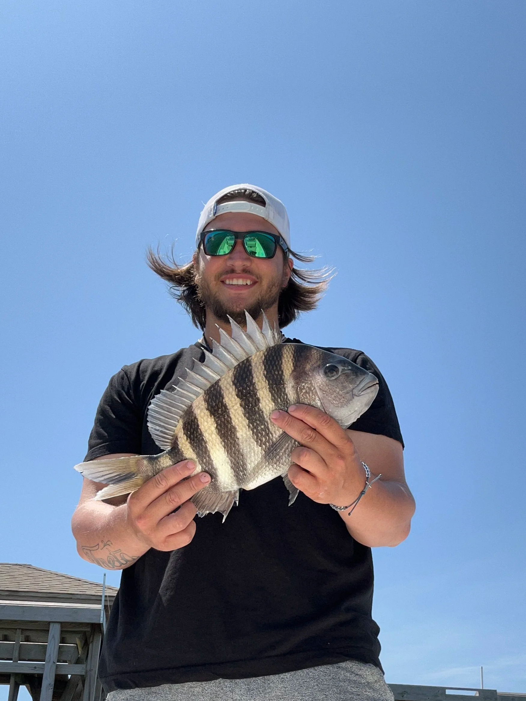 Man smiling, wearing sunglasses and a white cap, holding a large fish with vertical black and yellow stripes against a clear blue sky.