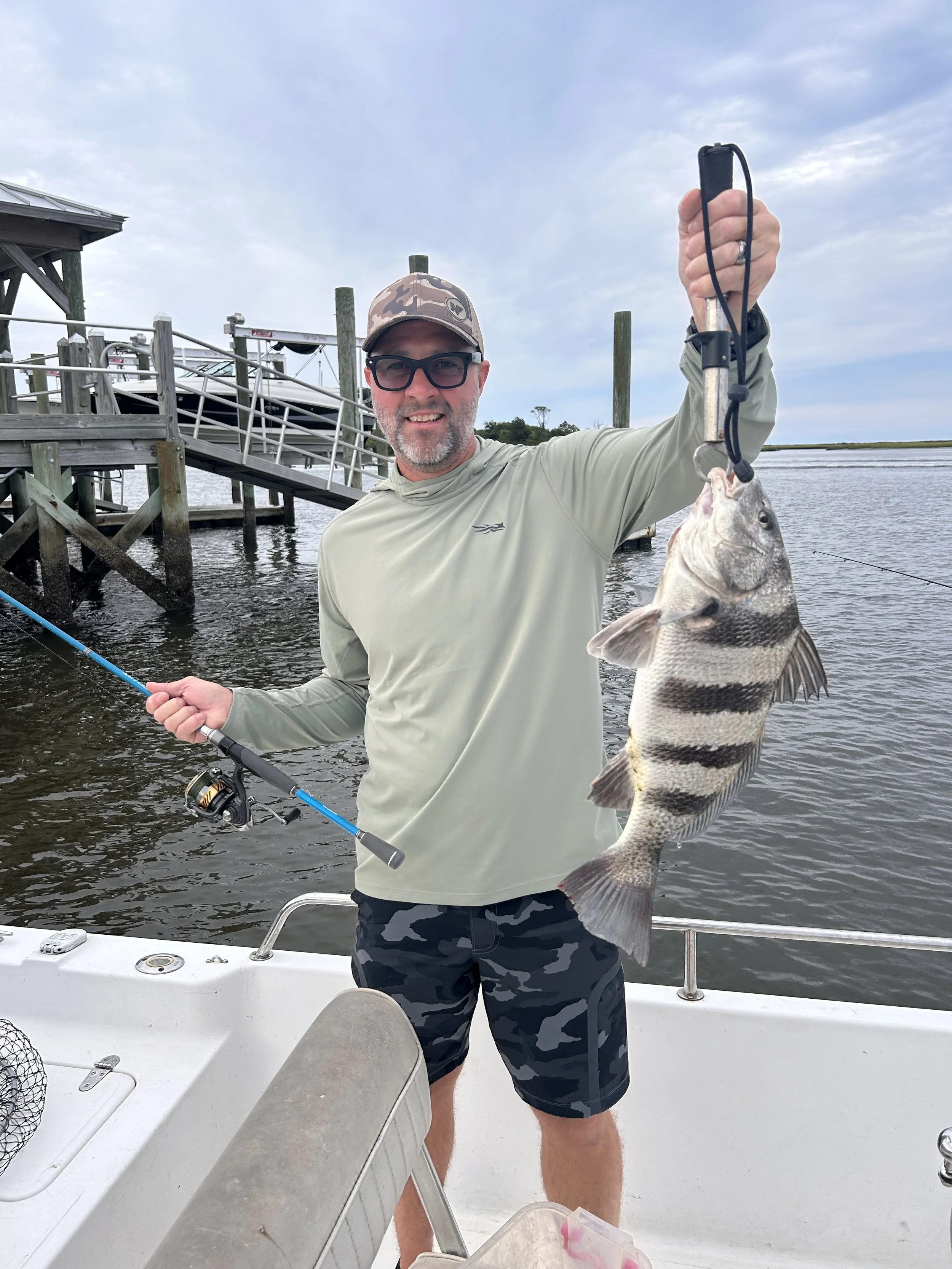 A man in sunglasses and a camouflage cap holding a striped fish on a boat, with a dock and water in the background.