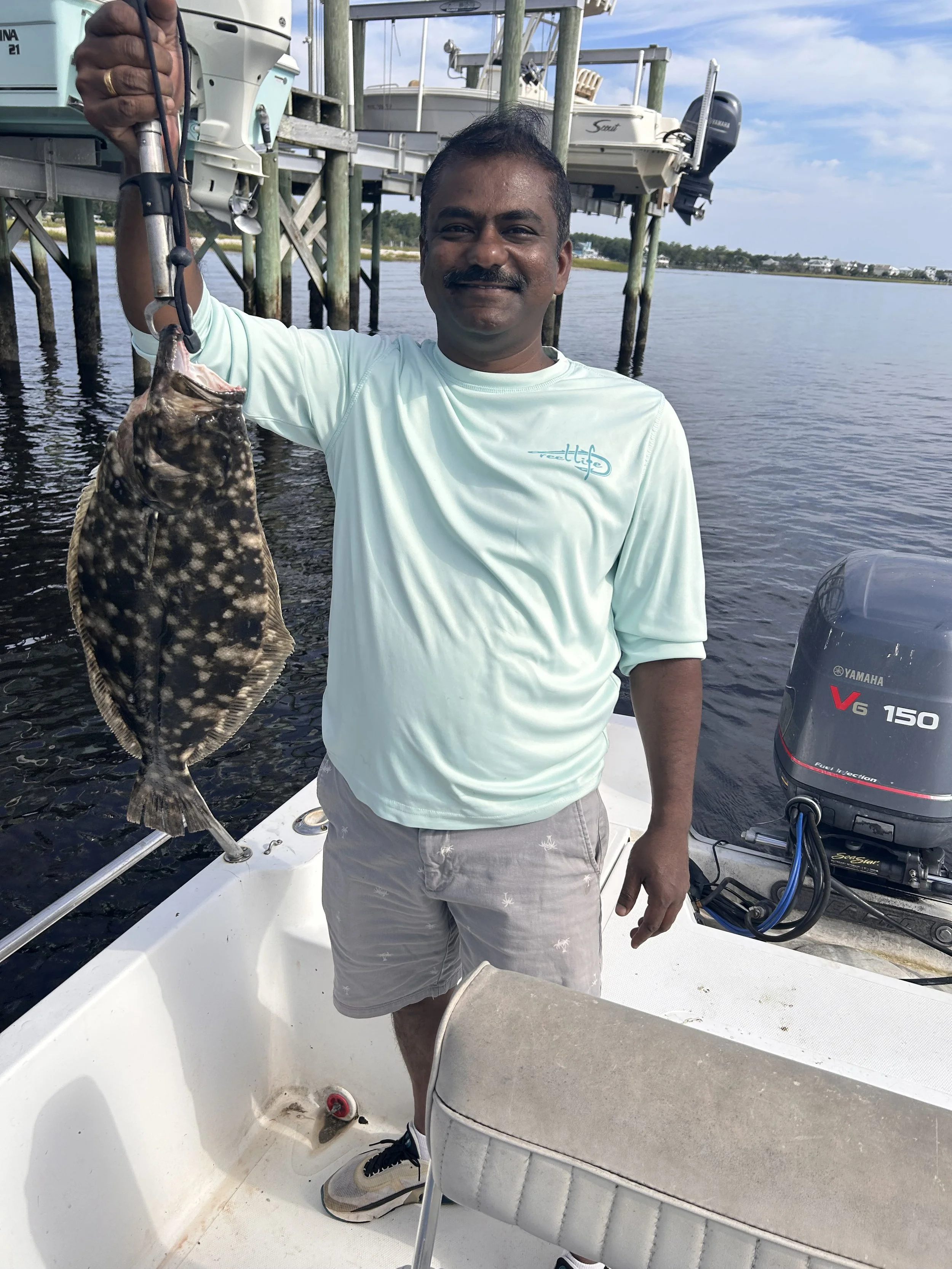 A man with a dark mustache and short hair standing on a boat dock, holding a fish with a fishing tool. The fish has dark body with white spots. The background shows water, a dock with boats, and a partly cloudy sky.