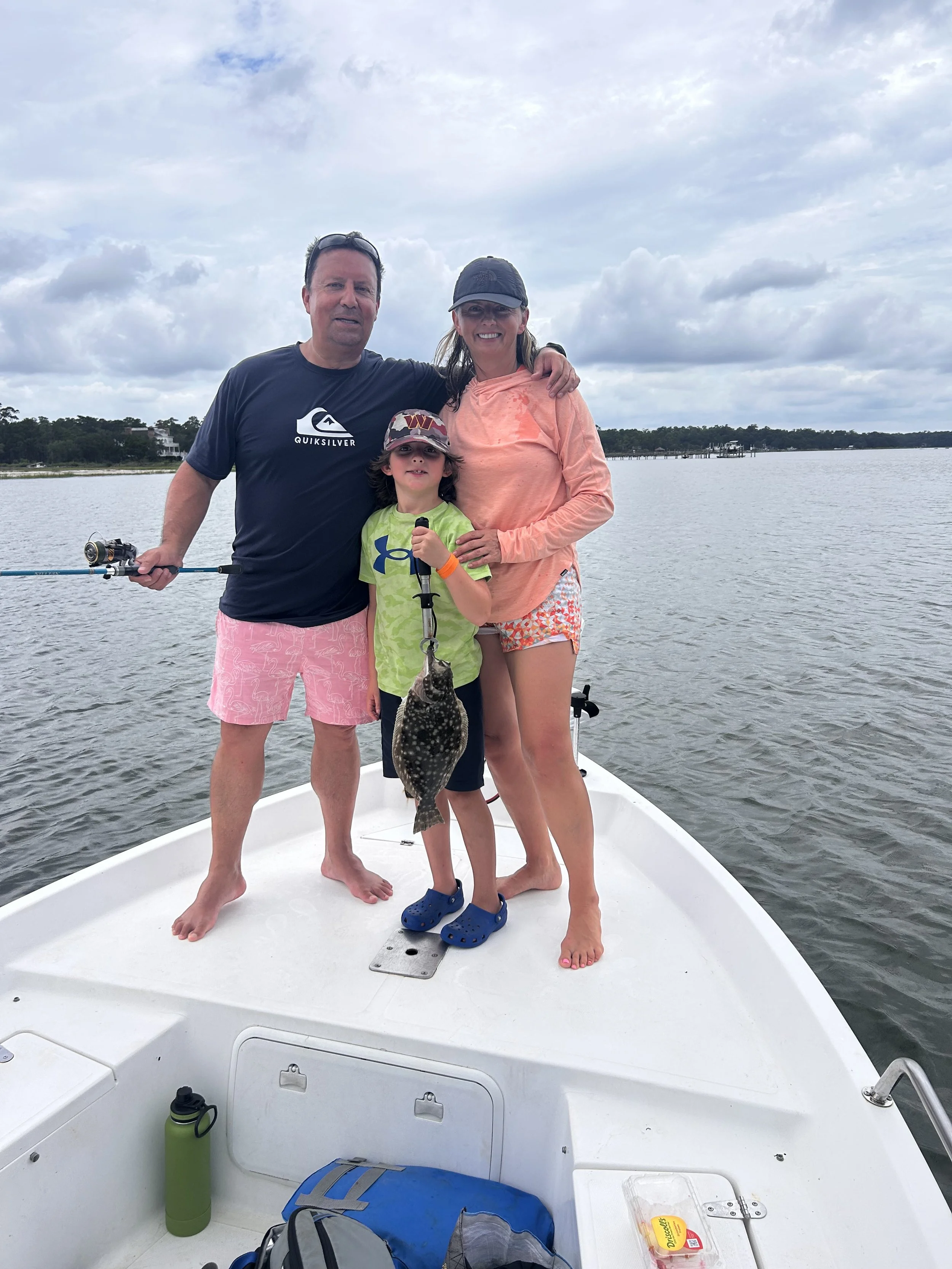 Family on a boat, with a boy holding a fish, in a lake or river under a cloudy sky.