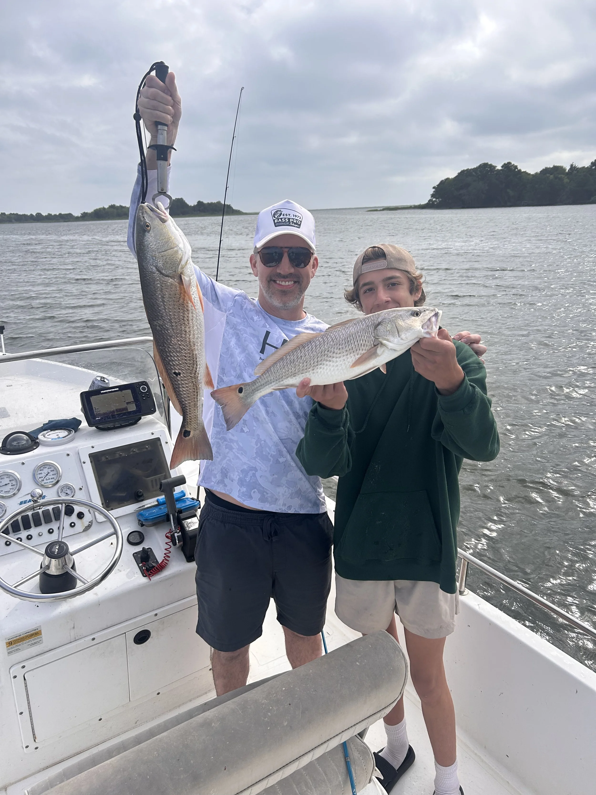 A man and a boy on a boat holding freshly caught fish, smiling at the camera, with water and cloudy sky in the background.