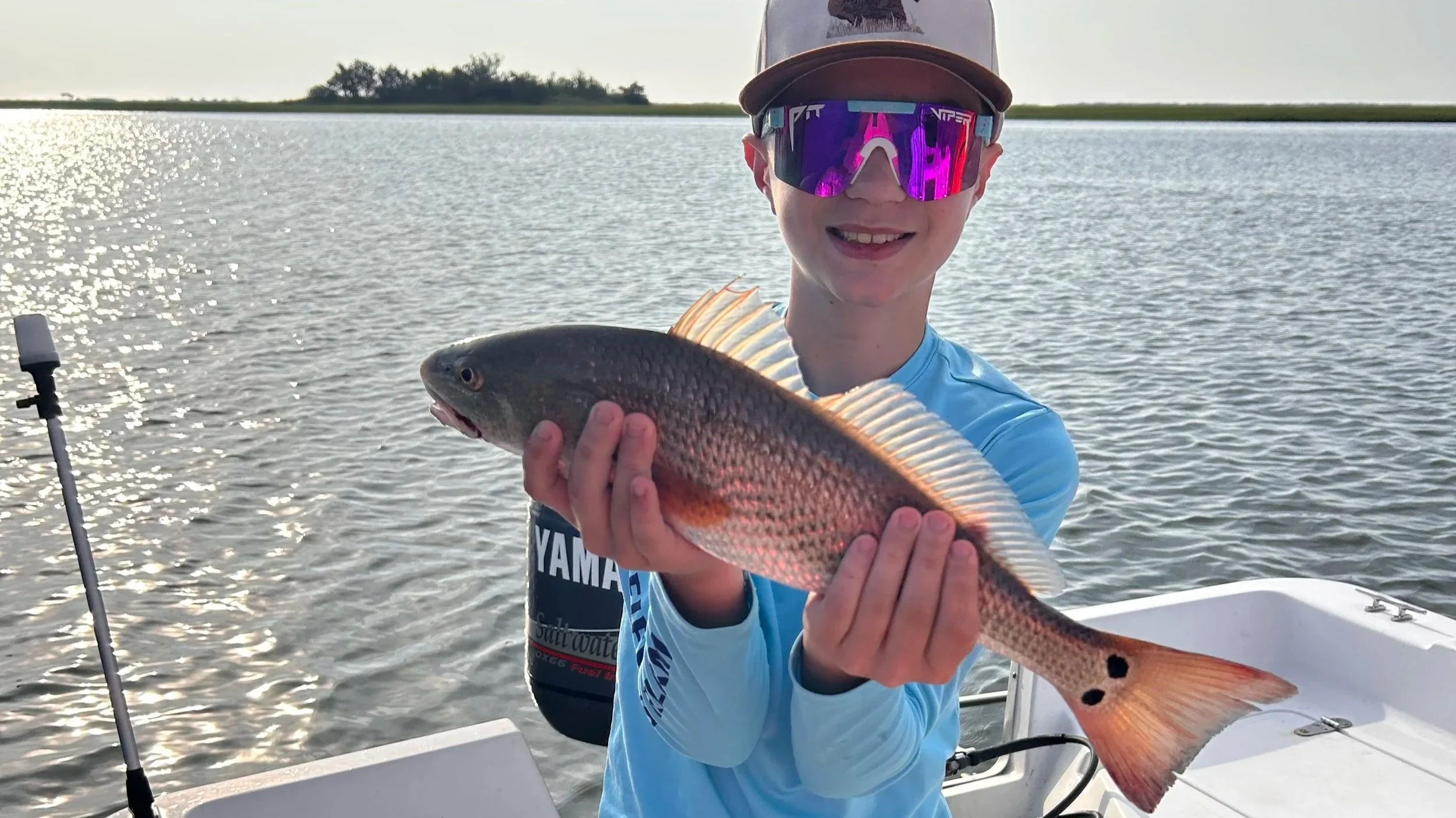 A smiling boy wearing a cap and sunglasses holding a fish with a multicolored body and two black spots on its tail, standing on a boat on a body of water during sunset.