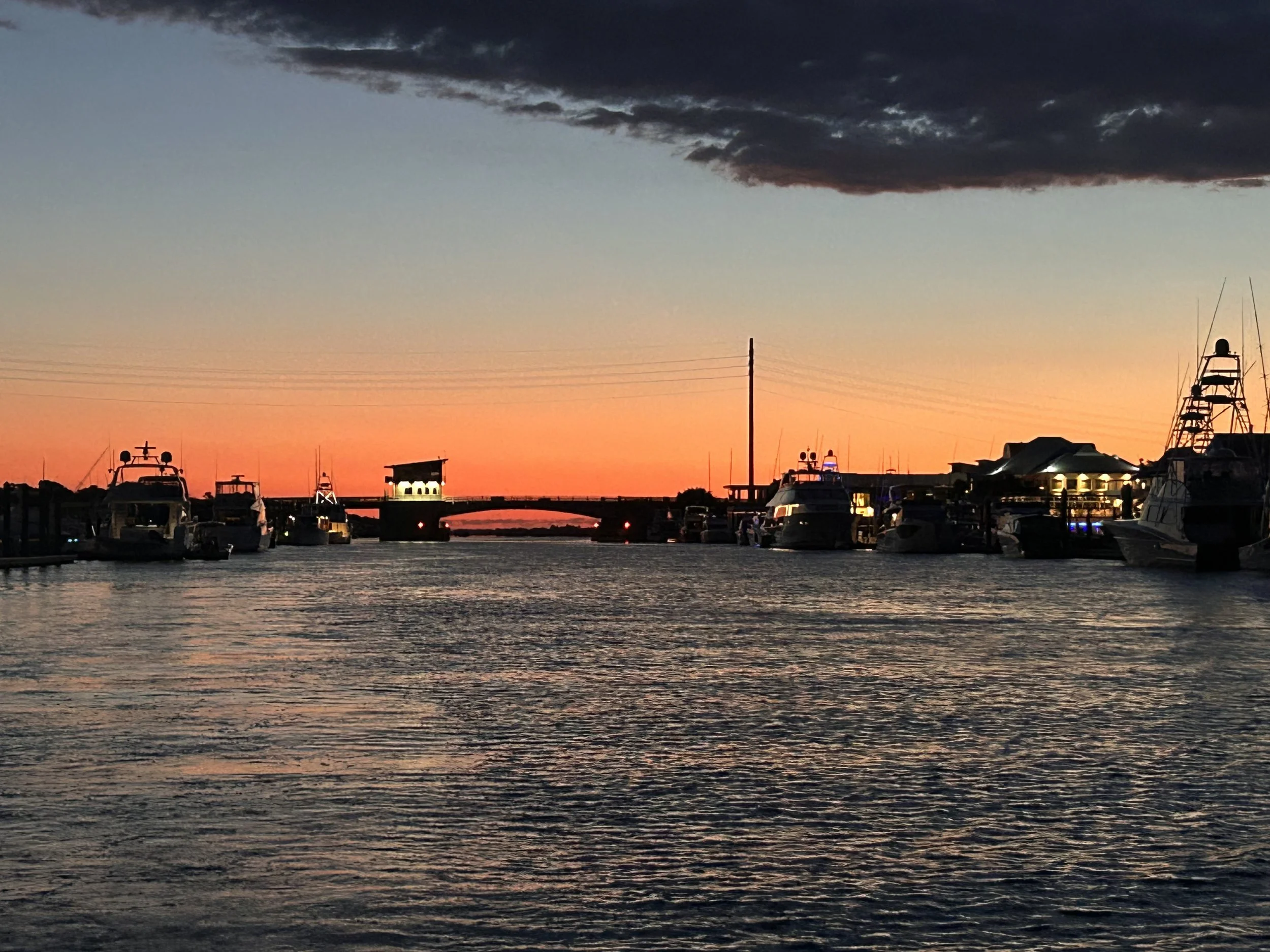 Boats docked at a marina during sunset, with a pink and orange sky and a bridge in the background.