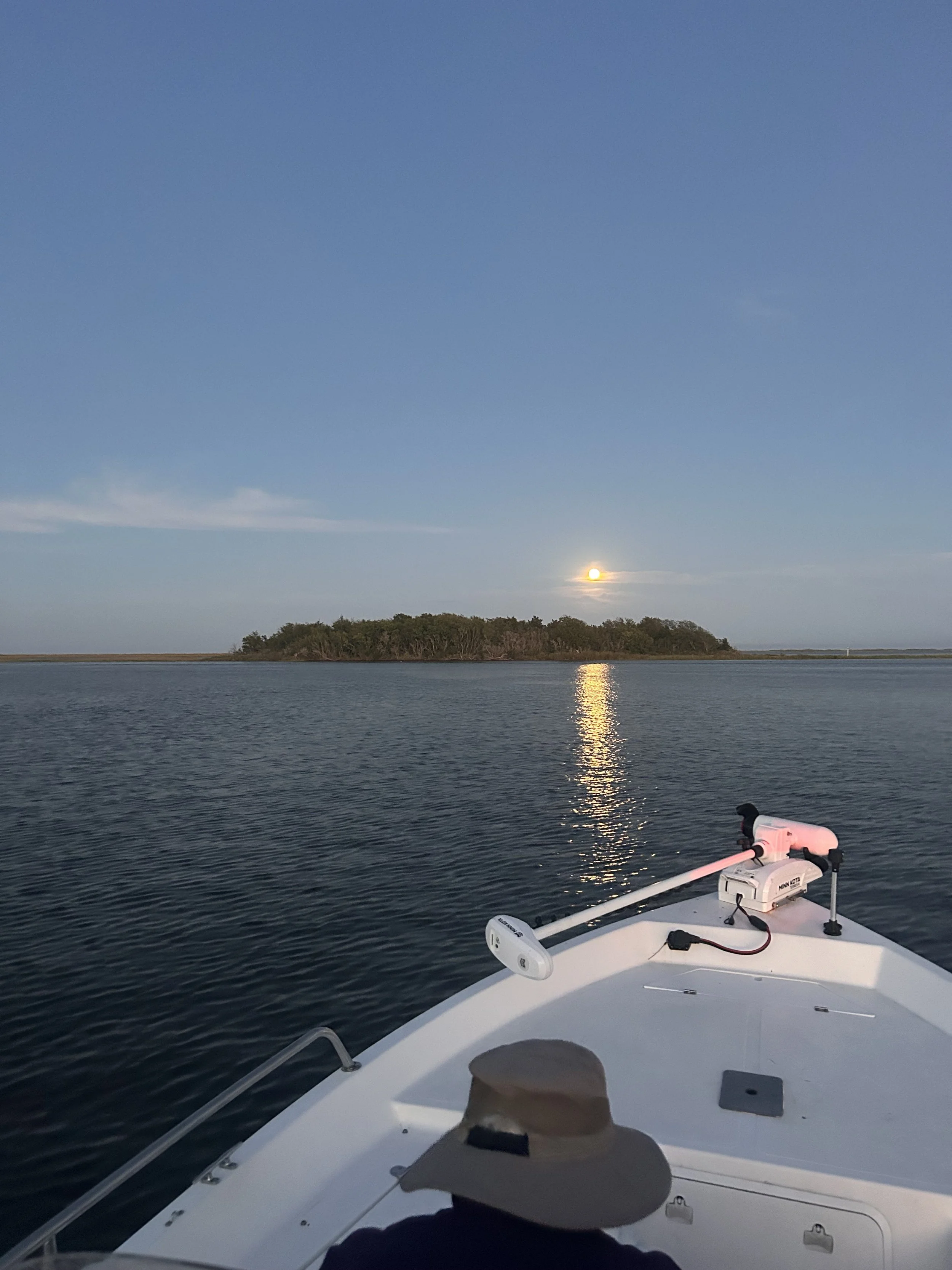 View from a boat on water during sunset or sunrise, with an island in the distance and a person wearing a hat in the lower part of the boat.