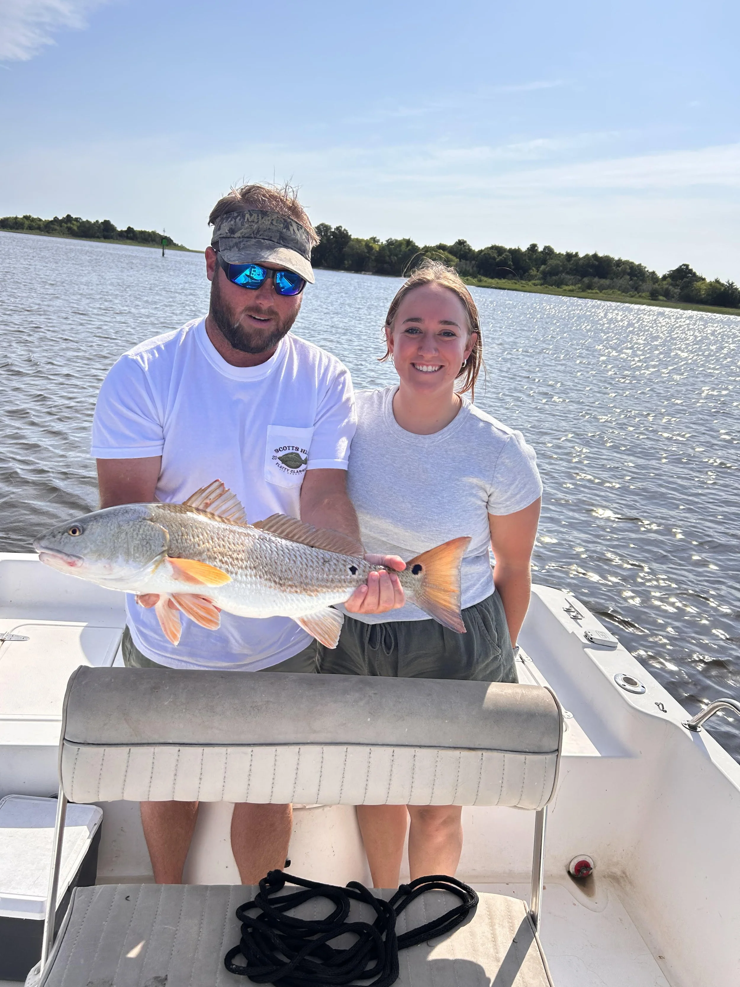 A man and a woman on a boat holding a fish they caught, with water and trees in the background on a sunny day.