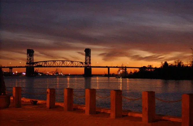Sunset over a river with a bridge and industrial area in the background, viewed from a promenade with orange bollards.