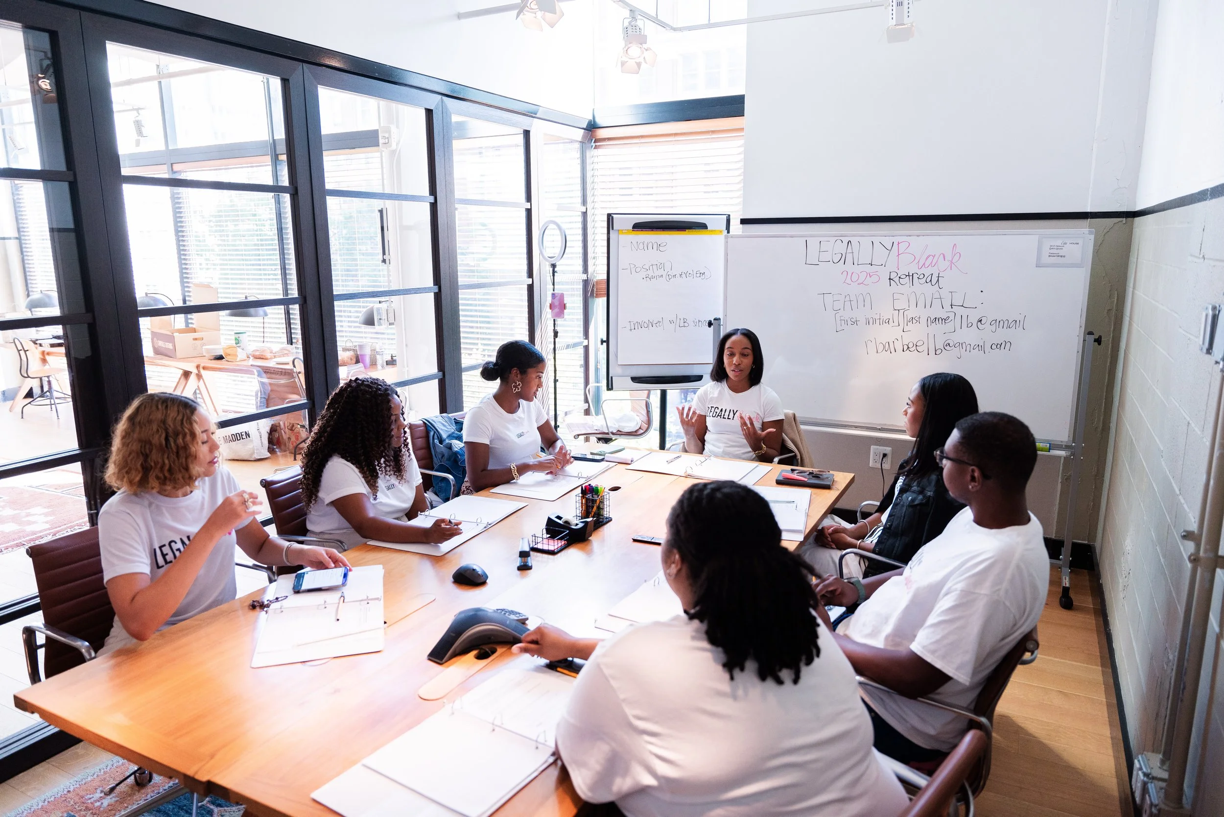 A group of women gathered around a conference table in a bright room with large windows, participating in a meeting or workshop. A woman at the front is speaking, with a whiteboard behind her showing notes and contact information.