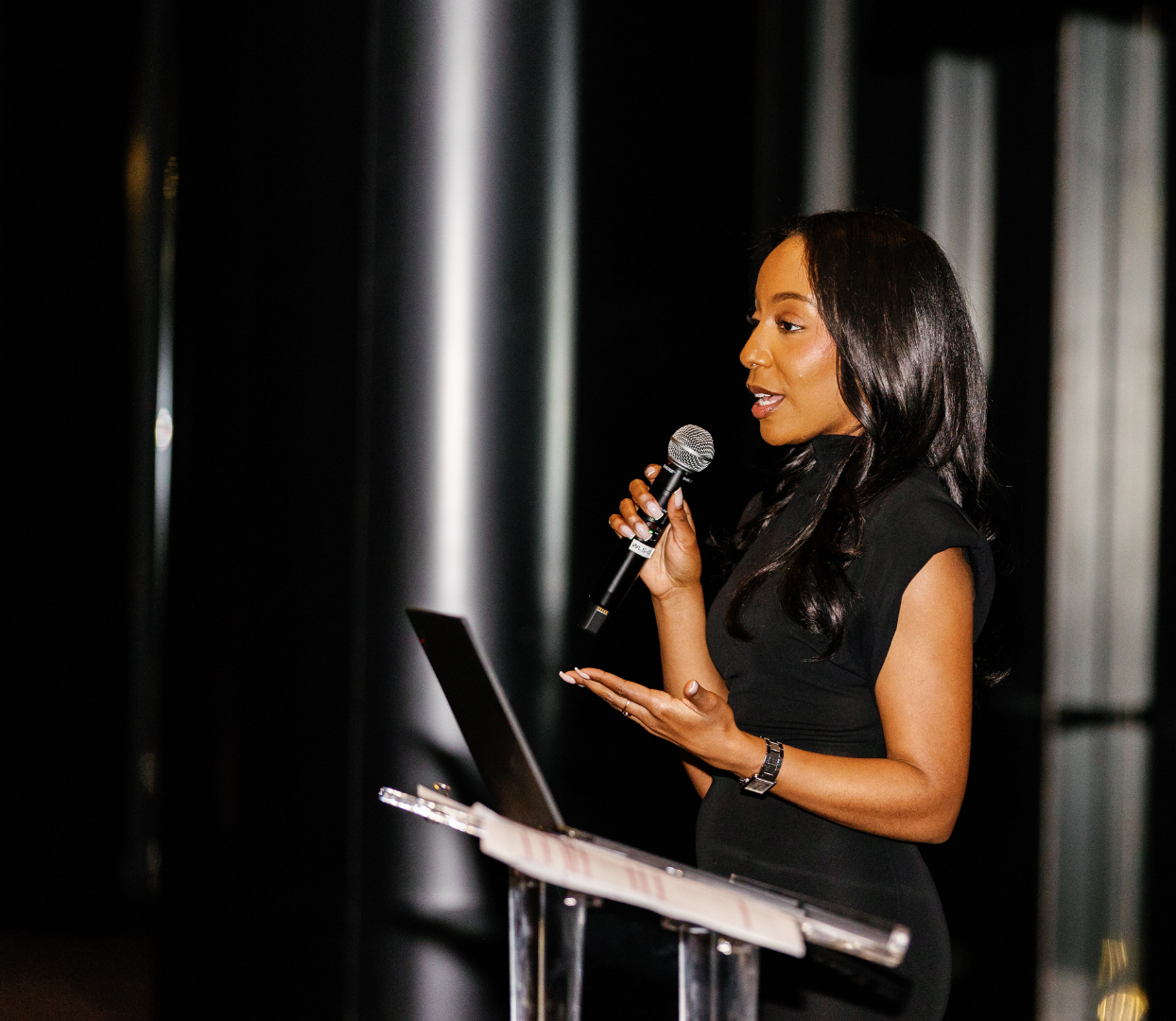 A woman with long dark hair in a black dress speaking into a microphone at a podium with a laptop.