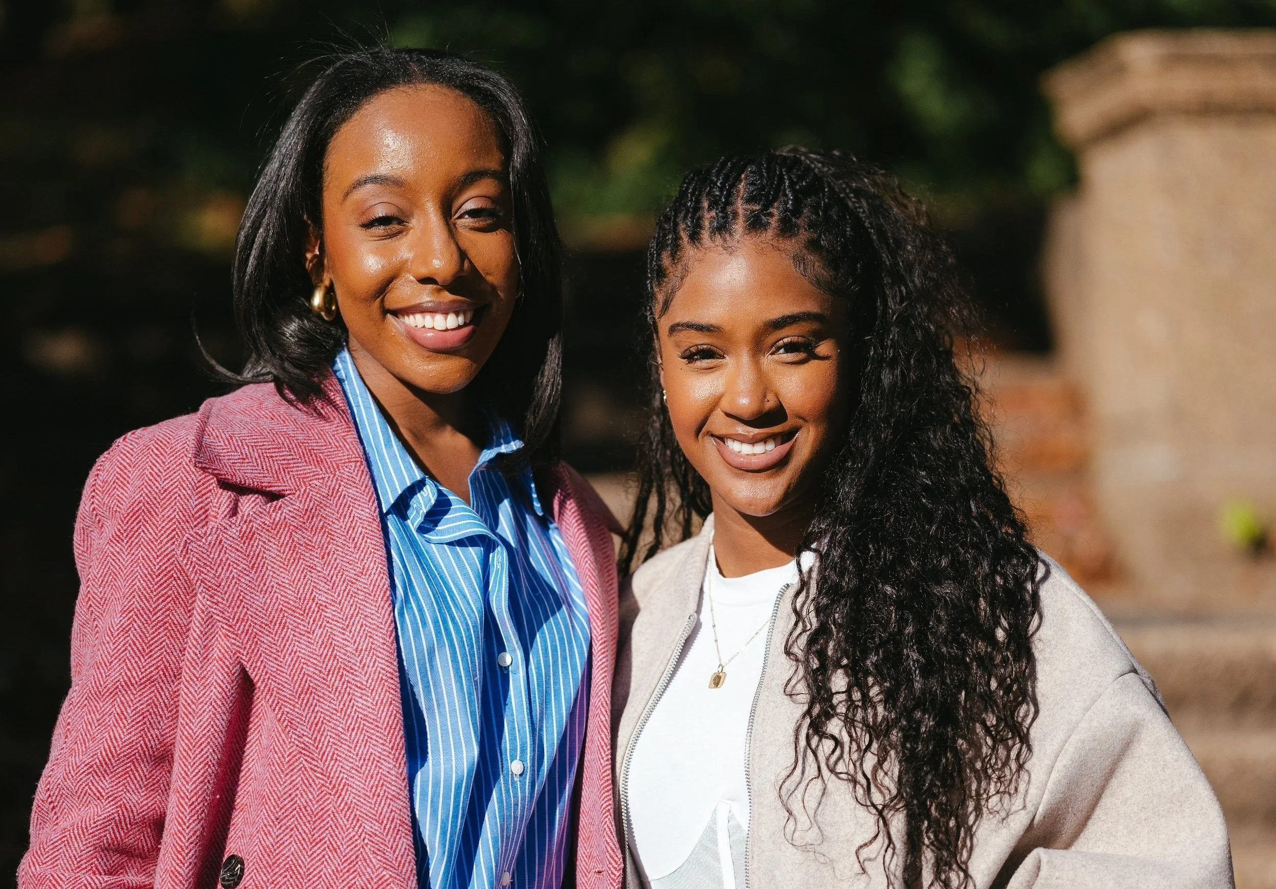 Two women smiling outdoors, standing close together with a blurred background of trees and a stone structure.