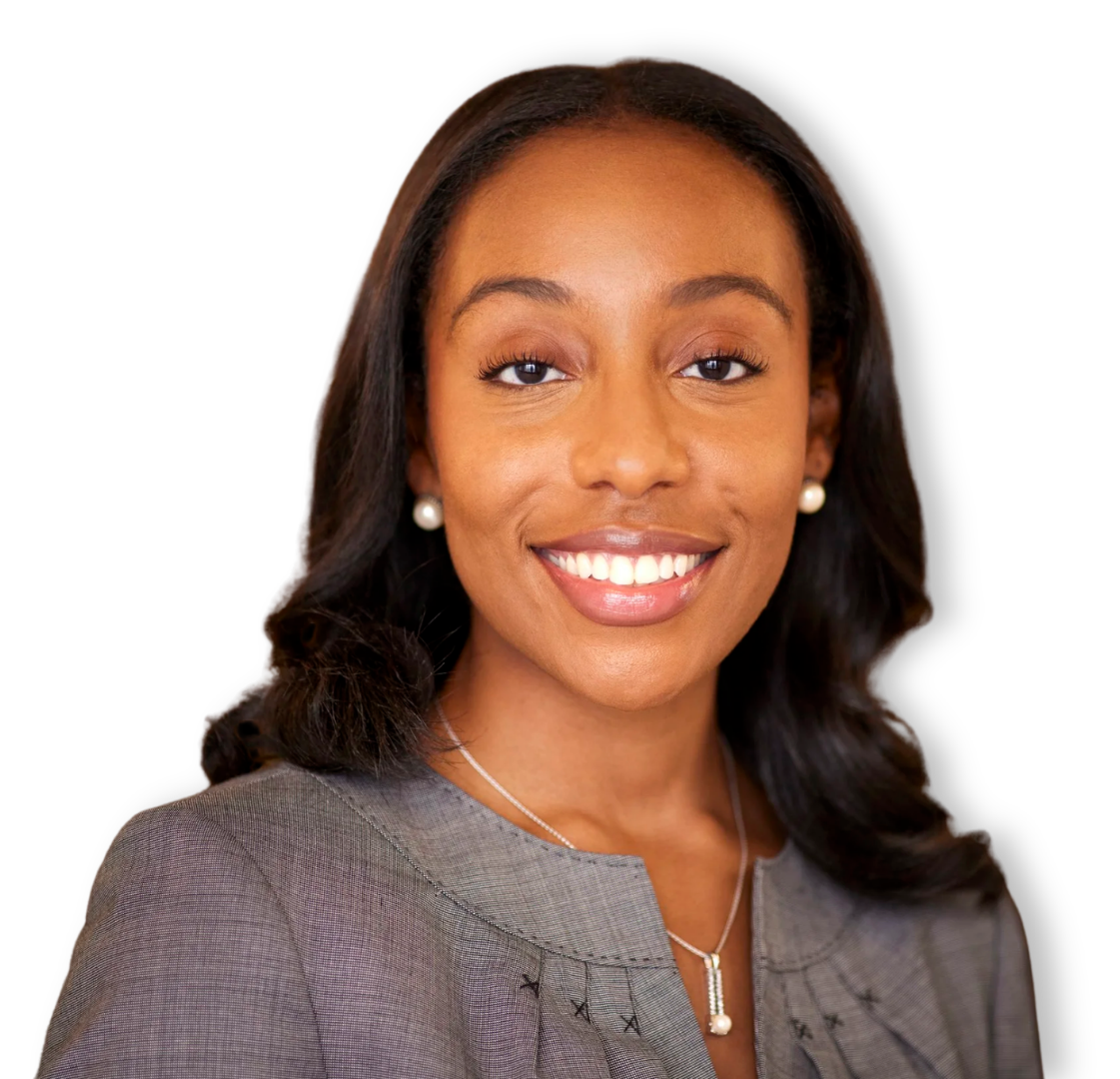 A woman with dark hair, wearing pearl earrings, a gray blazer, and a pearl pendant necklace, smiling at the camera against a black background.