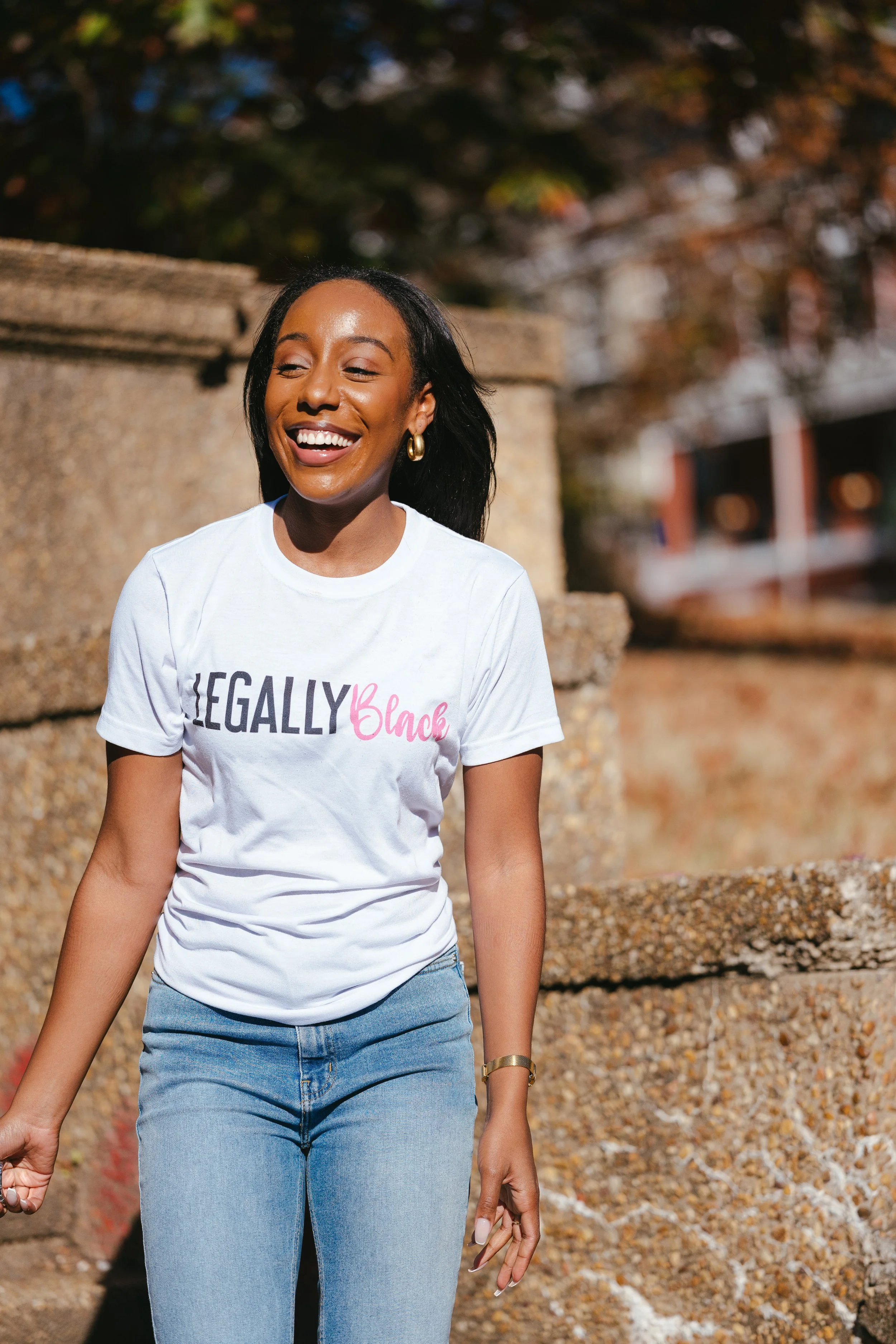A woman smiling outdoors near a stone wall, wearing a white t-shirt with the words 'Legally Black' and light blue jeans.