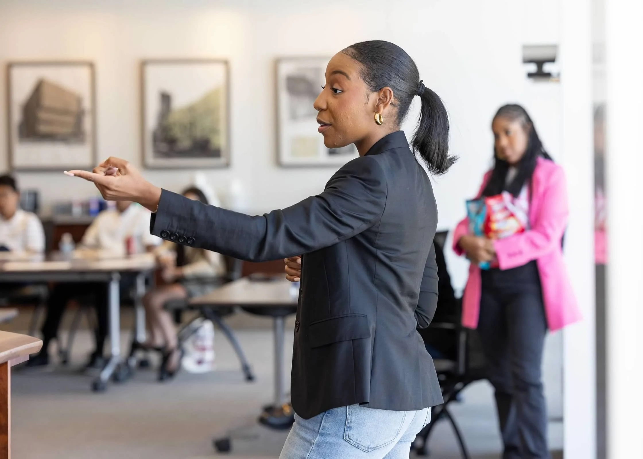 A woman in a black blazer and light blue jeans speaking or presenting in a meeting room with artworks on the wall, while another woman in a pink jacket and holding a pink bag looks on in the background.