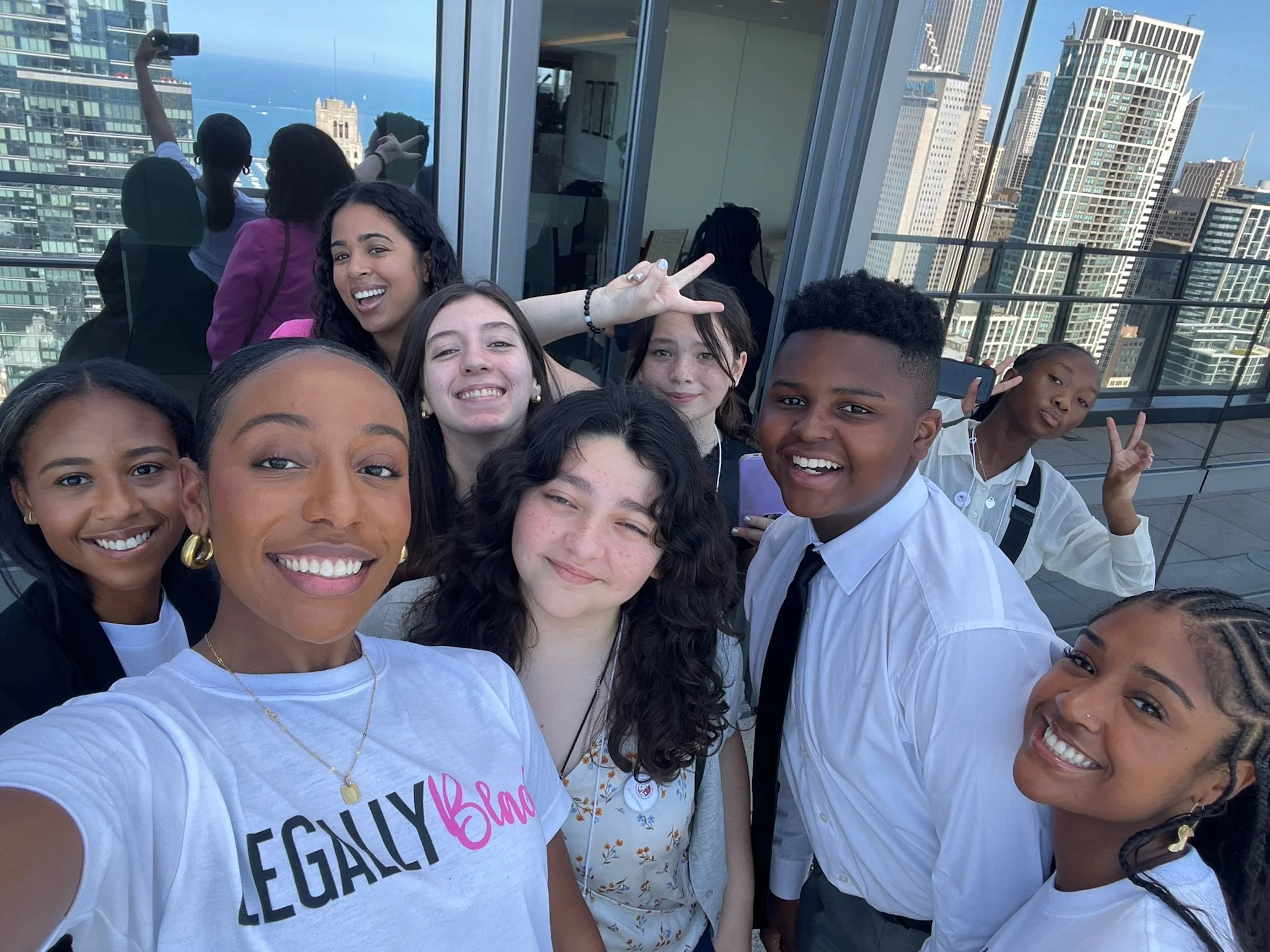 A group of young people taking a selfie on a rooftop with a city skyline in the background, smiling and making peace signs.