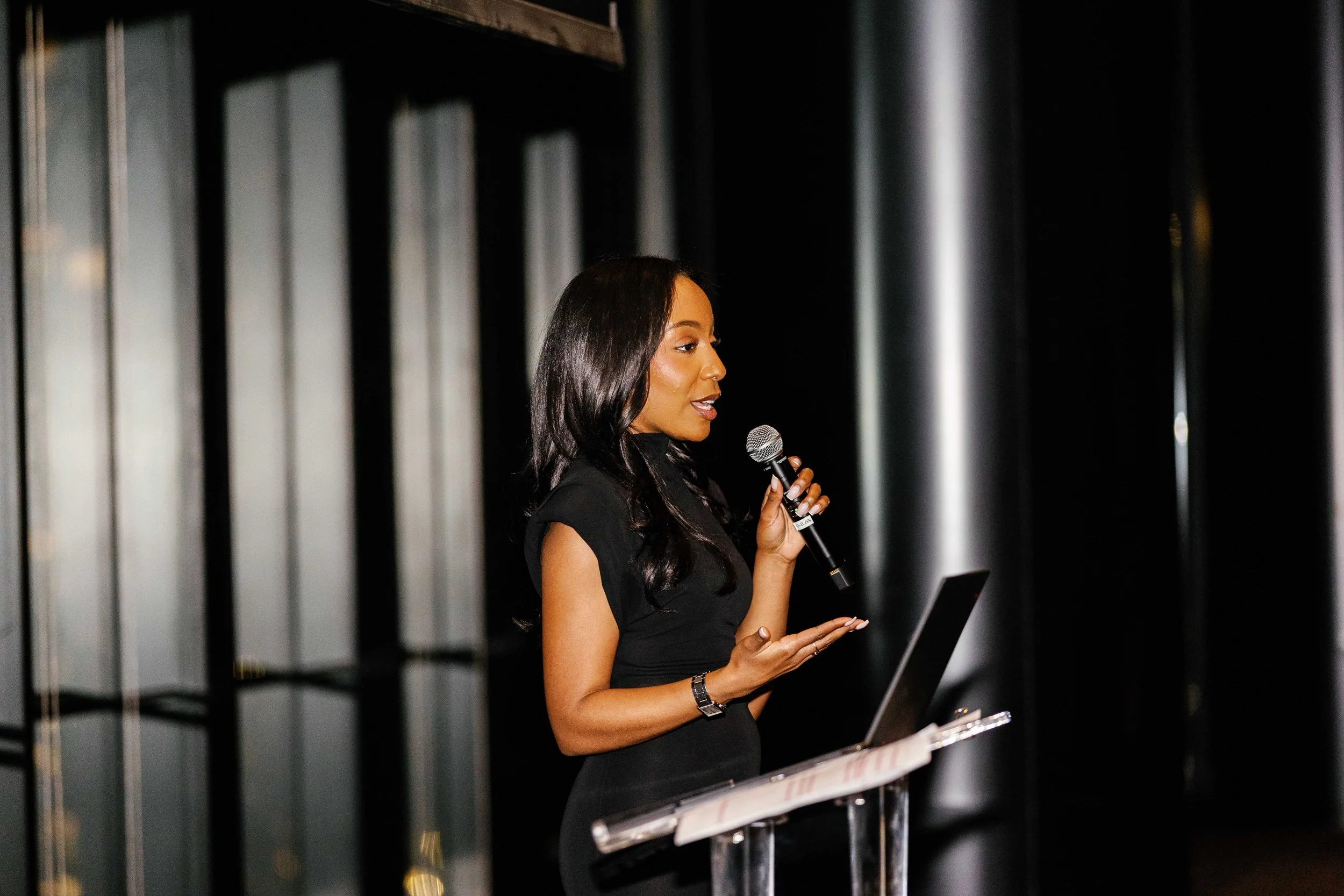 A woman with long black hair, wearing a black dress, holding a microphone, standing in front of a dark background with vertical metallic bars, possibly giving a presentation or speech.
