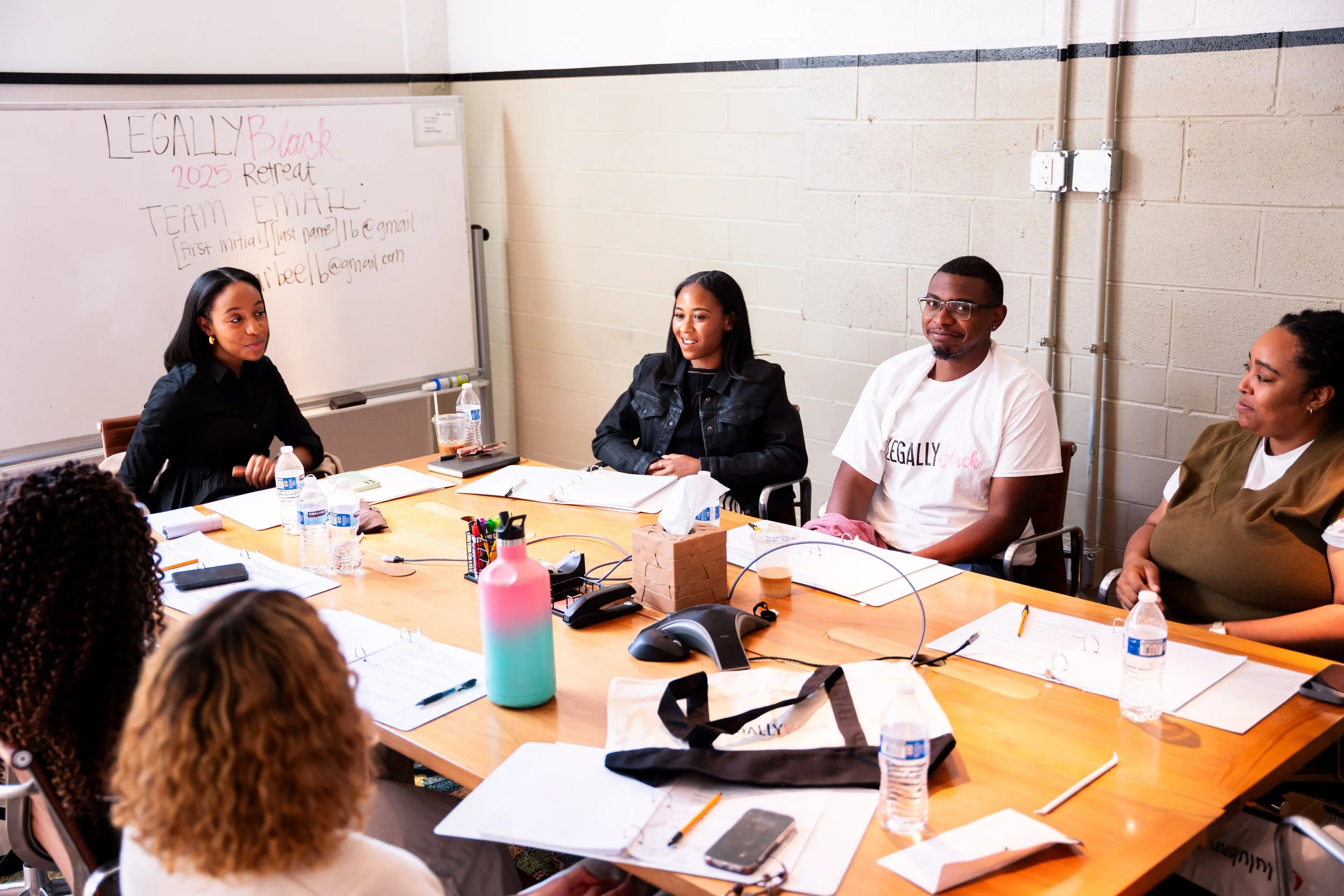A group of people sitting around a large wooden table in a room with white walls. There are water bottles, notebooks, pens, and a pink and teal water bottle on the table. A whiteboard in the background has handwritten notes and the words "Legally Black 2025 Retreat".