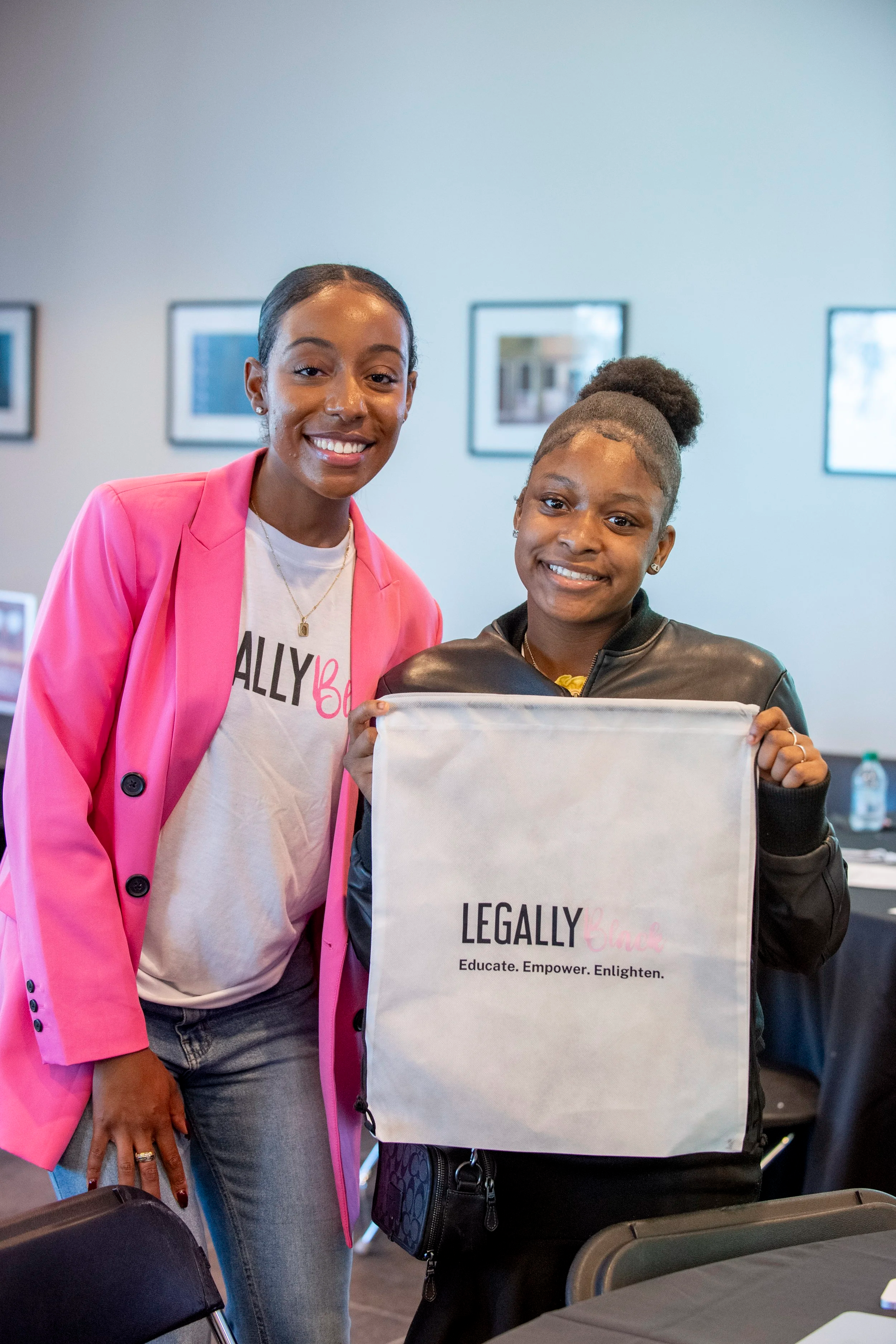 Two women standing together indoors, one holding a white tote bag with the words "Legally Black" and the subtitle "Educate. Empower. Enlighten." The woman on the left is wearing a bright pink blazer over a white t-shirt with text, and the woman on the right has curly hair styled in an updo, wearing a black leather jacket. They are smiling at the camera.