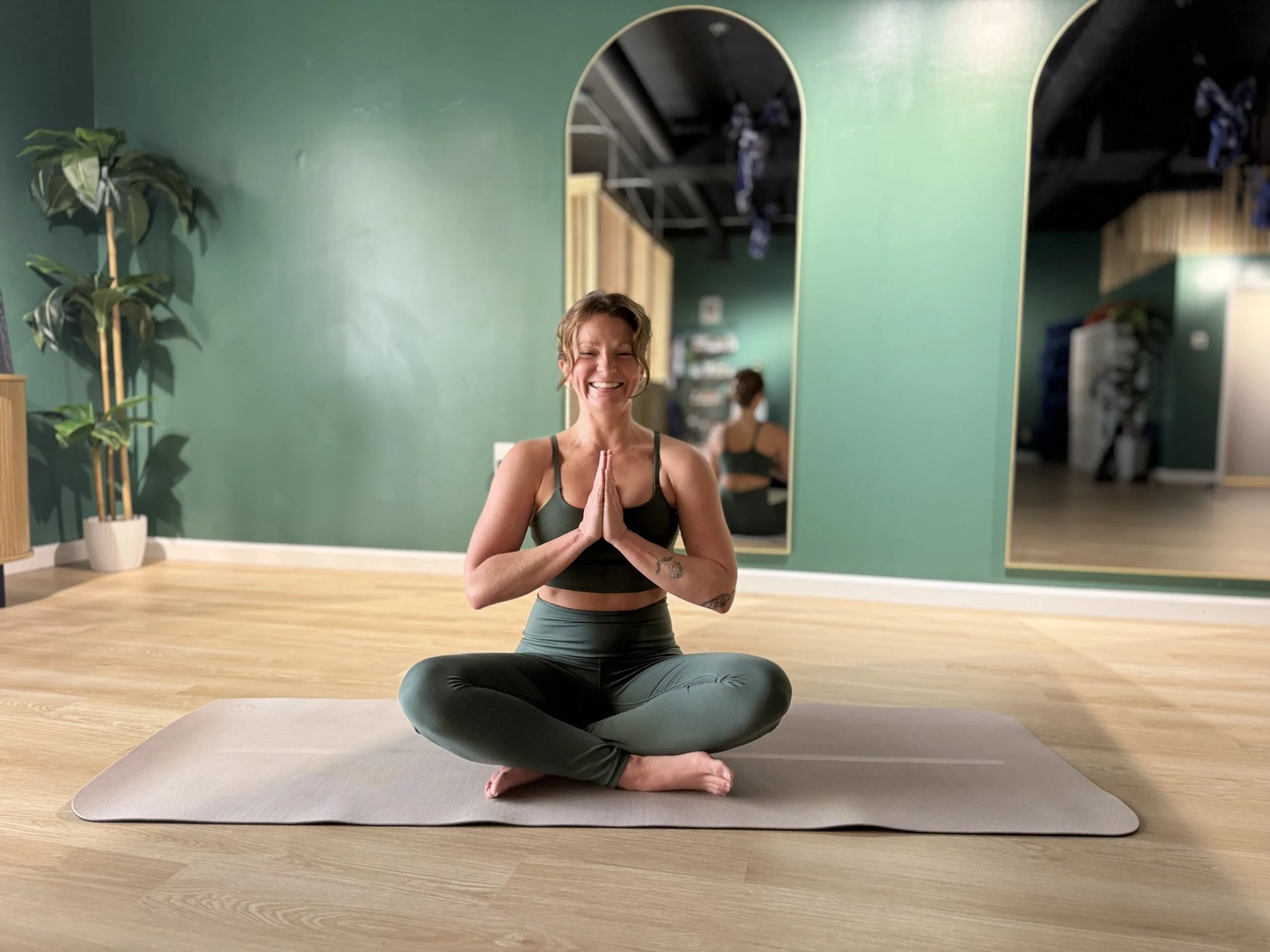 A woman practicing yoga indoors, sitting cross-legged on a yoga mat, with her hands in a prayer position and smiling, in a room with green walls, mirrors, and indoor plants.