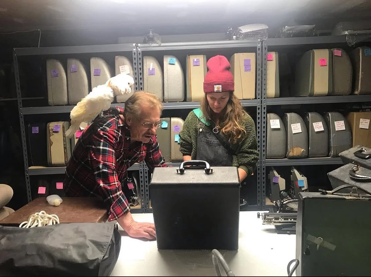 Glenwood Springs office supply owner and a woman examining typewriters on a table in storage room with shelves of vintage suitcases and bags in background; a white 55-year-old Moluccan Cockatoo perched on the man's shoulder.
