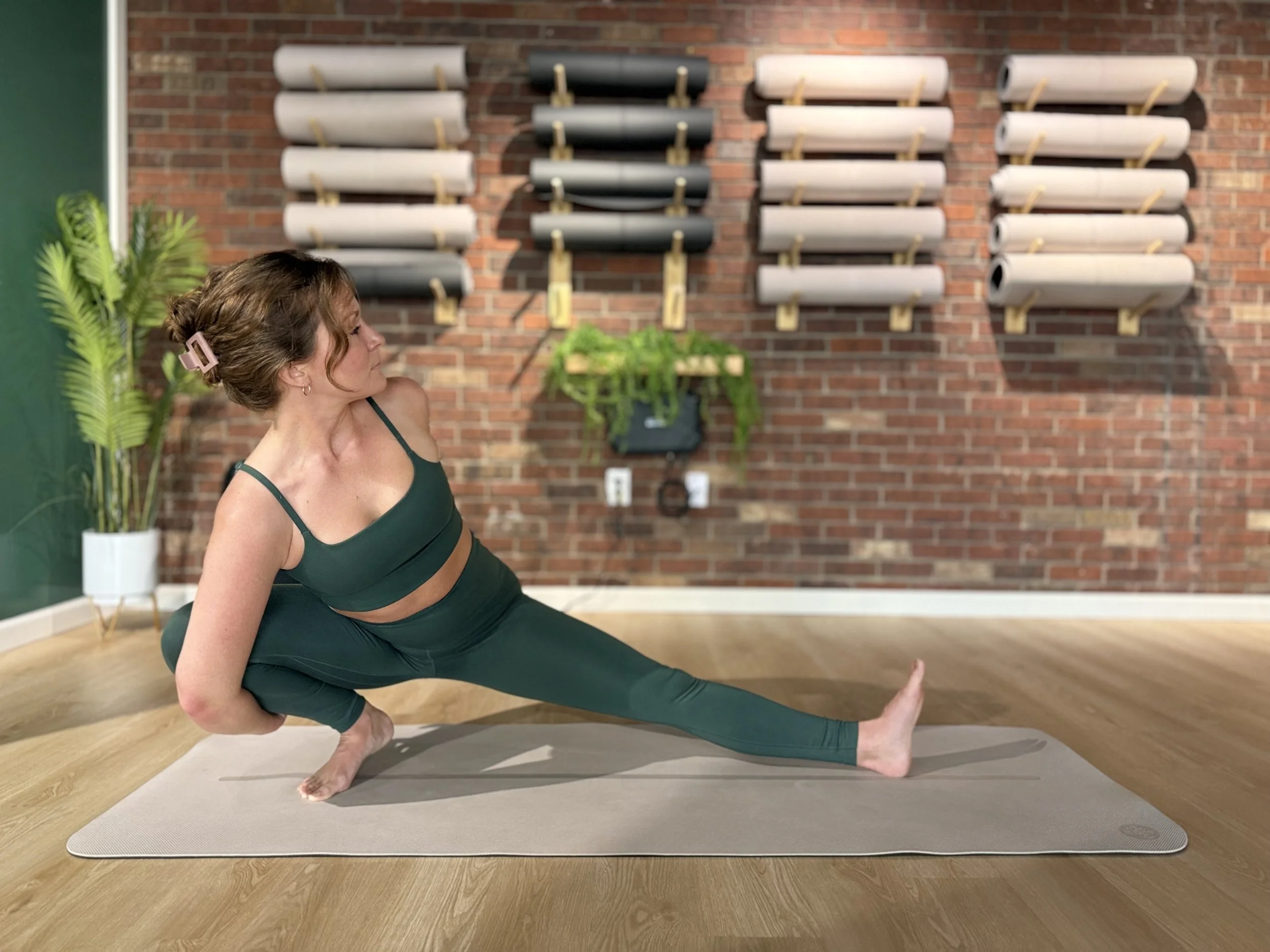 A woman in sportswear performing a yoga pose in a studio with brick walls, yoga mats, and plants.