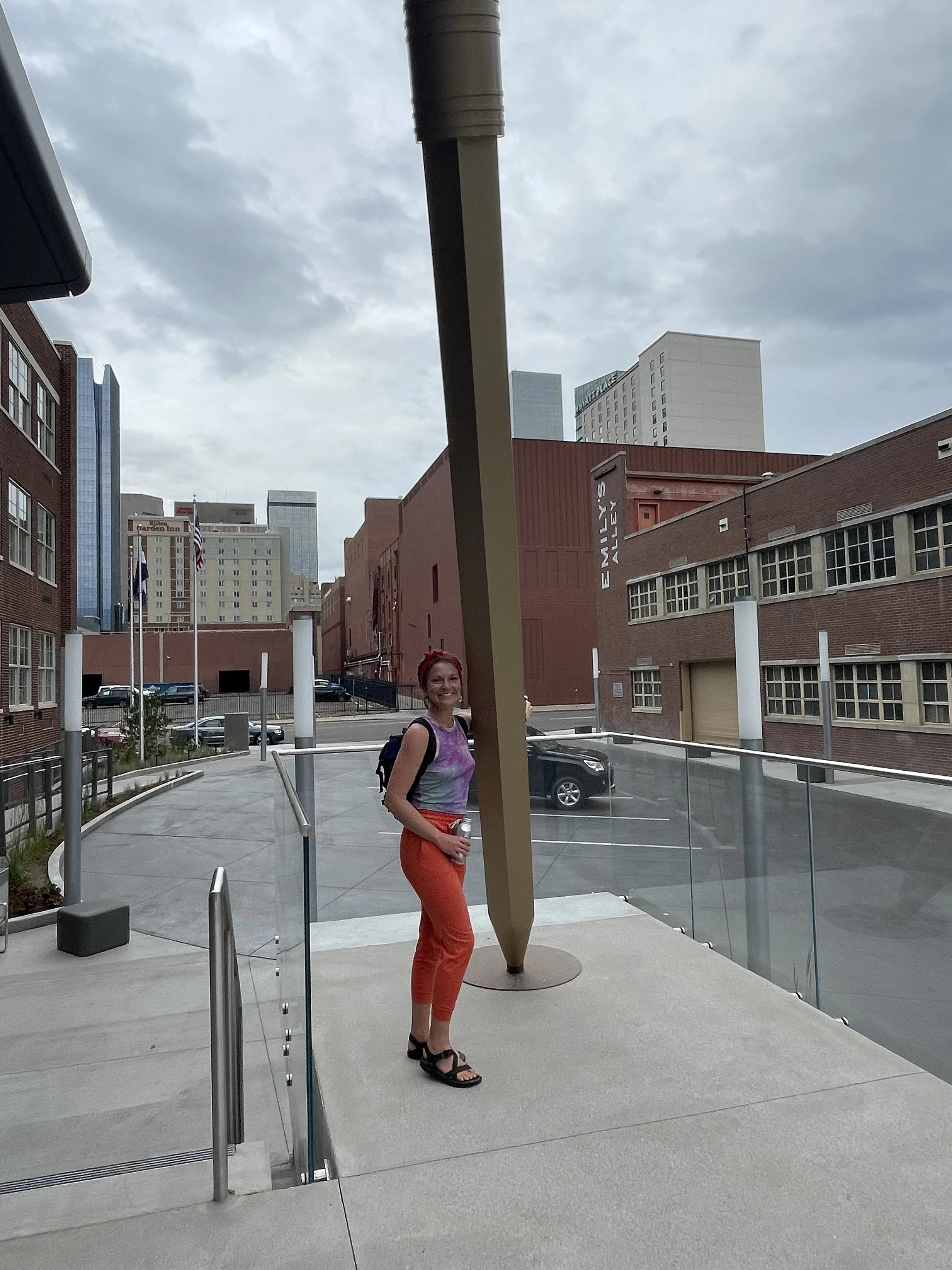 A woman with red hair, wearing a colorful sleeveless top and red pants, stands outdoors next to a large sculpture of a pencil. She is smiling and holding a water bottle. The background shows modern buildings and a cloudy sky.