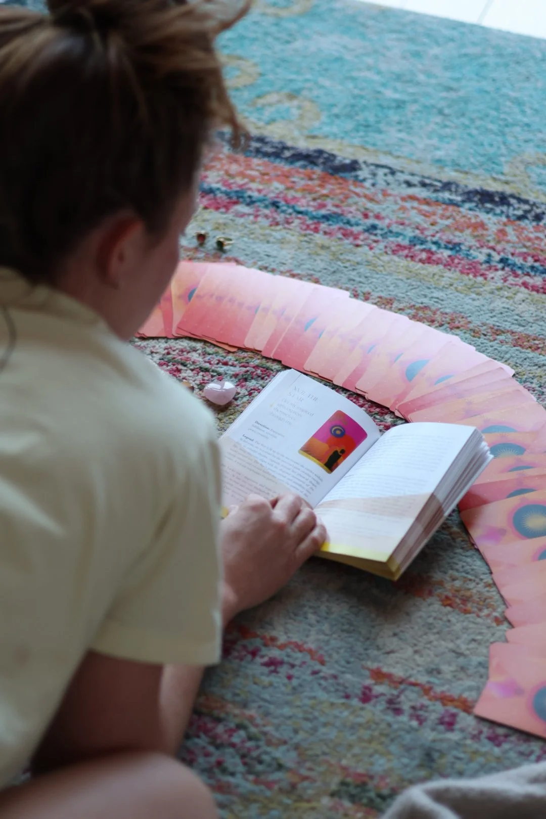 A woman on a colorful patterned carpet, looking at an open book with pink tarot cards splayed in a semi-circle.
