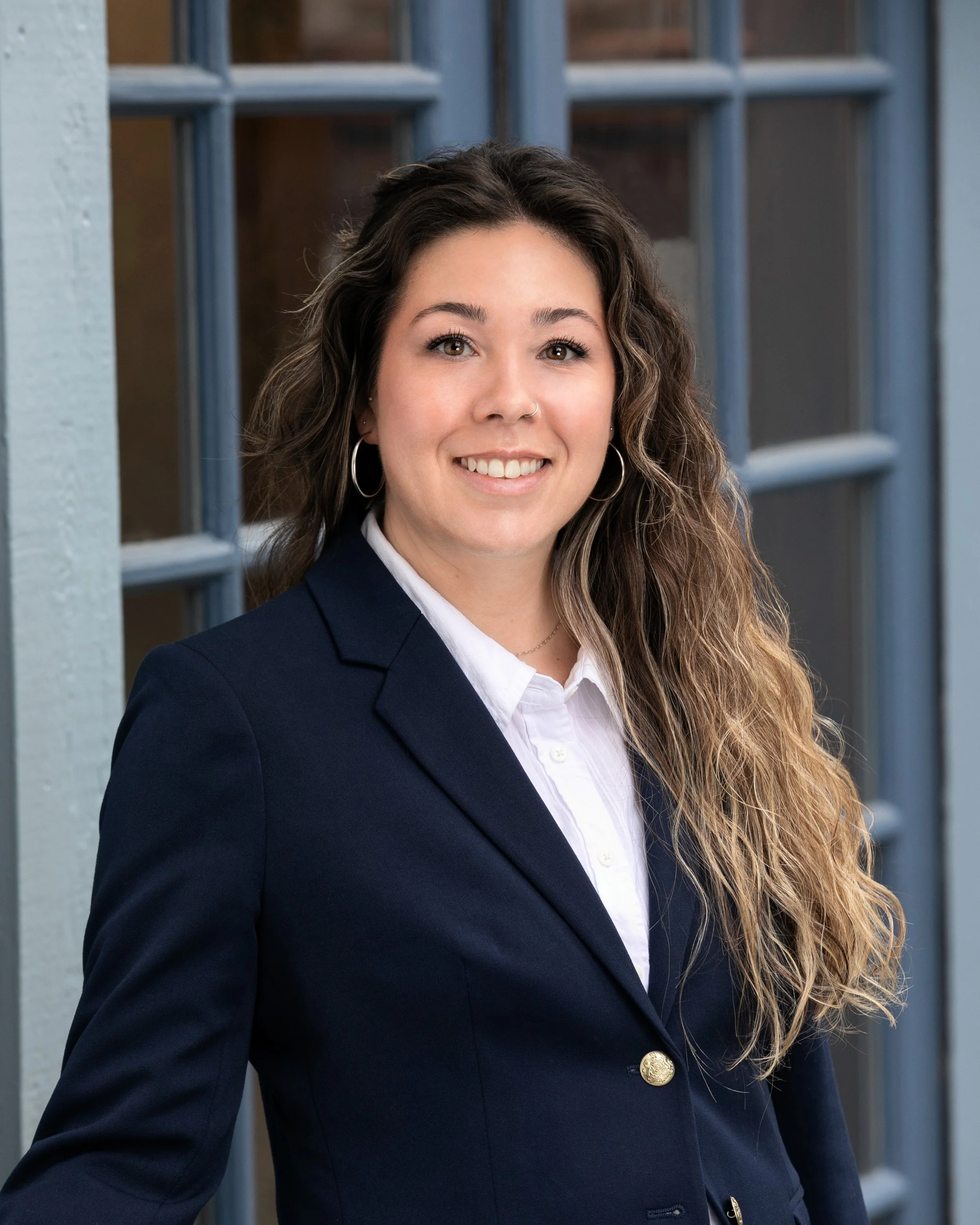A woman with long wavy hair, wearing a navy blazer and white shirt, standing outdoors in front of a blue window.