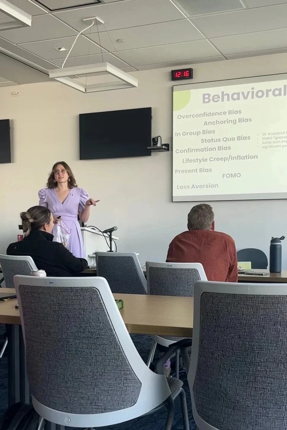 A woman in a lavender dress giving a presentation in a classroom. There are students seated at tables, and a slide titled 'Behavioral' with a list of biases is projected on the wall.