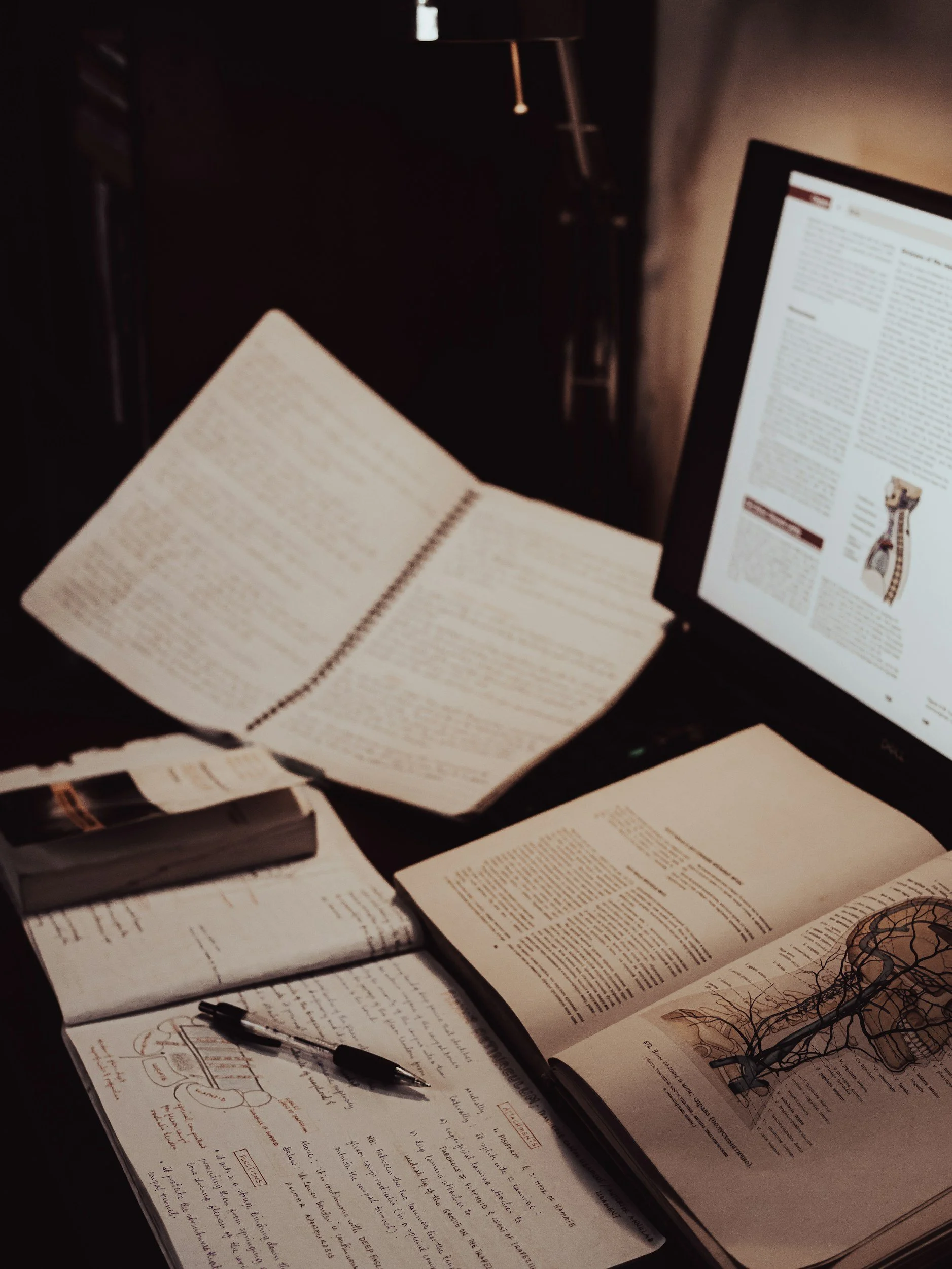 Study desk with open textbooks, papers, notebooks, a pen, and a computer monitor displaying a webpage, in a dimly lit room.