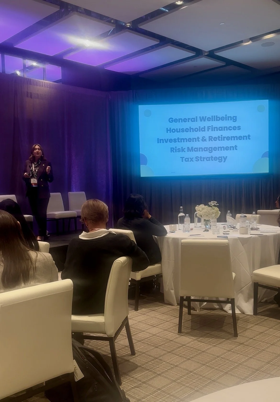 A woman giving a presentation at a conference with a slide displaying topics like General Wellbeing, Household Finances, Investment & Retirement, Risk Management, and Tax Strategy. There are seated attendees and a table with water bottles and flowers in the foreground.