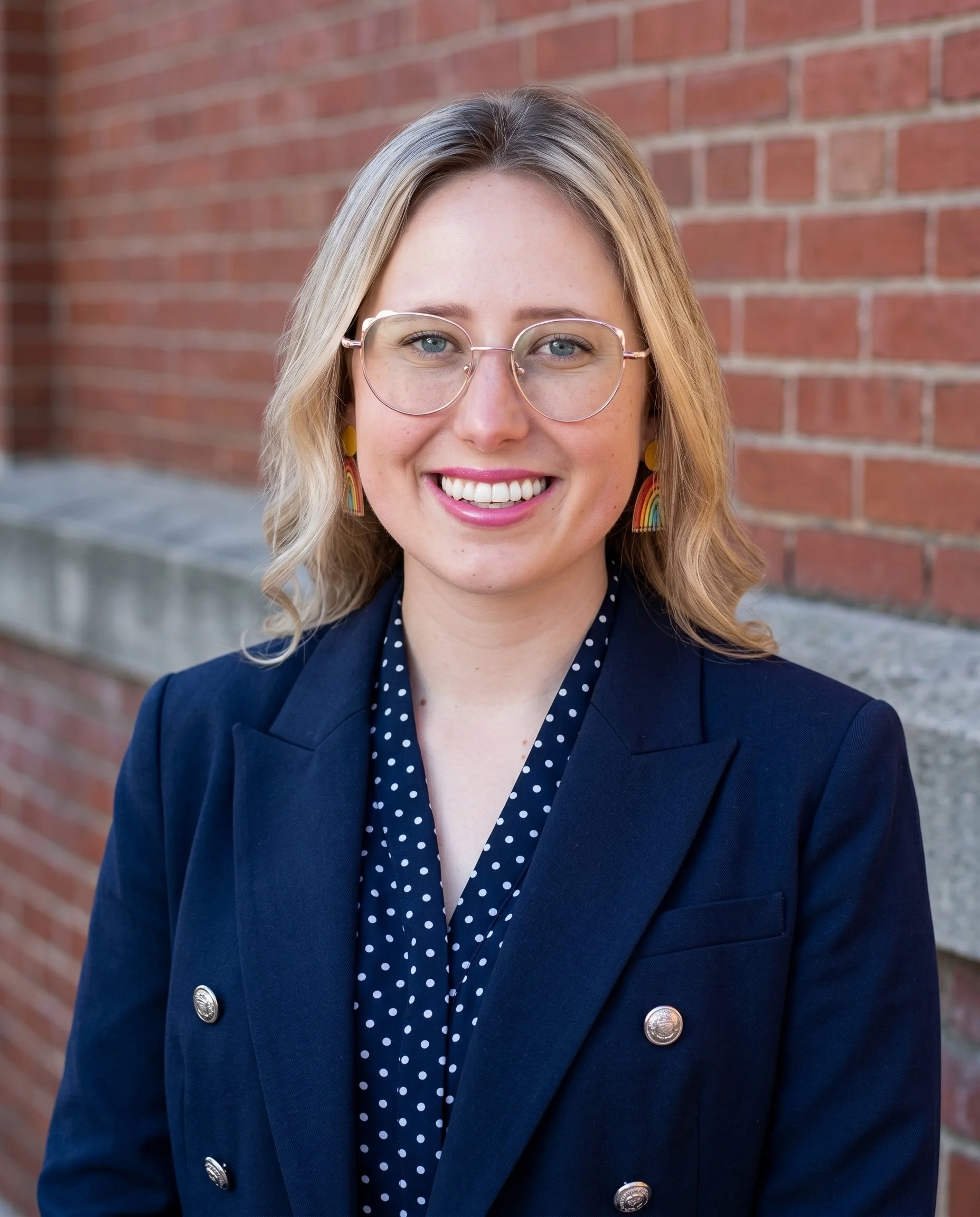Close-up of a woman with blonde hair, glasses, rainbow earrings, wearing a navy blazer and polka dot blouse, smiling in front of a brick wall.