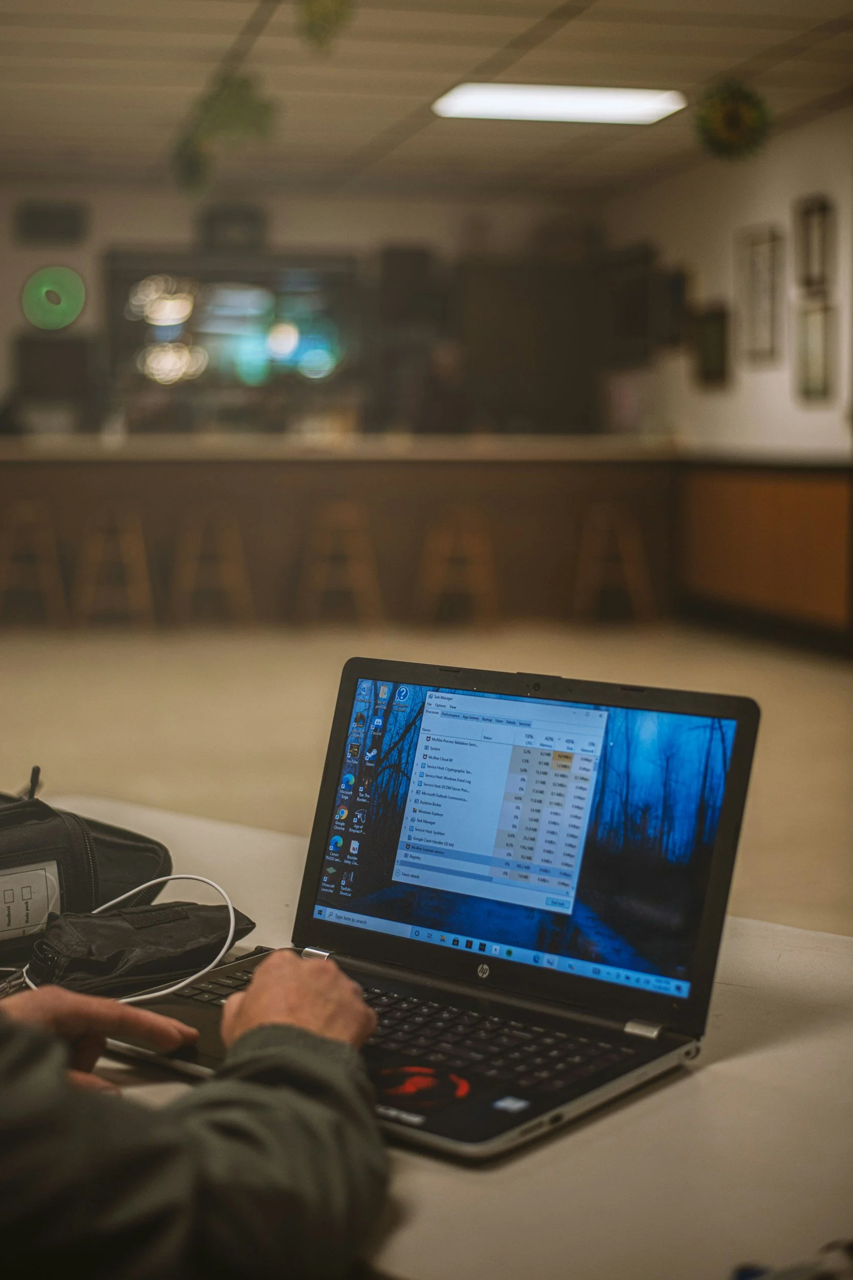 Person working on a laptop in a room with a bar counter in the background. The laptop screen displays a software window with data, and the person is using a mouse.
