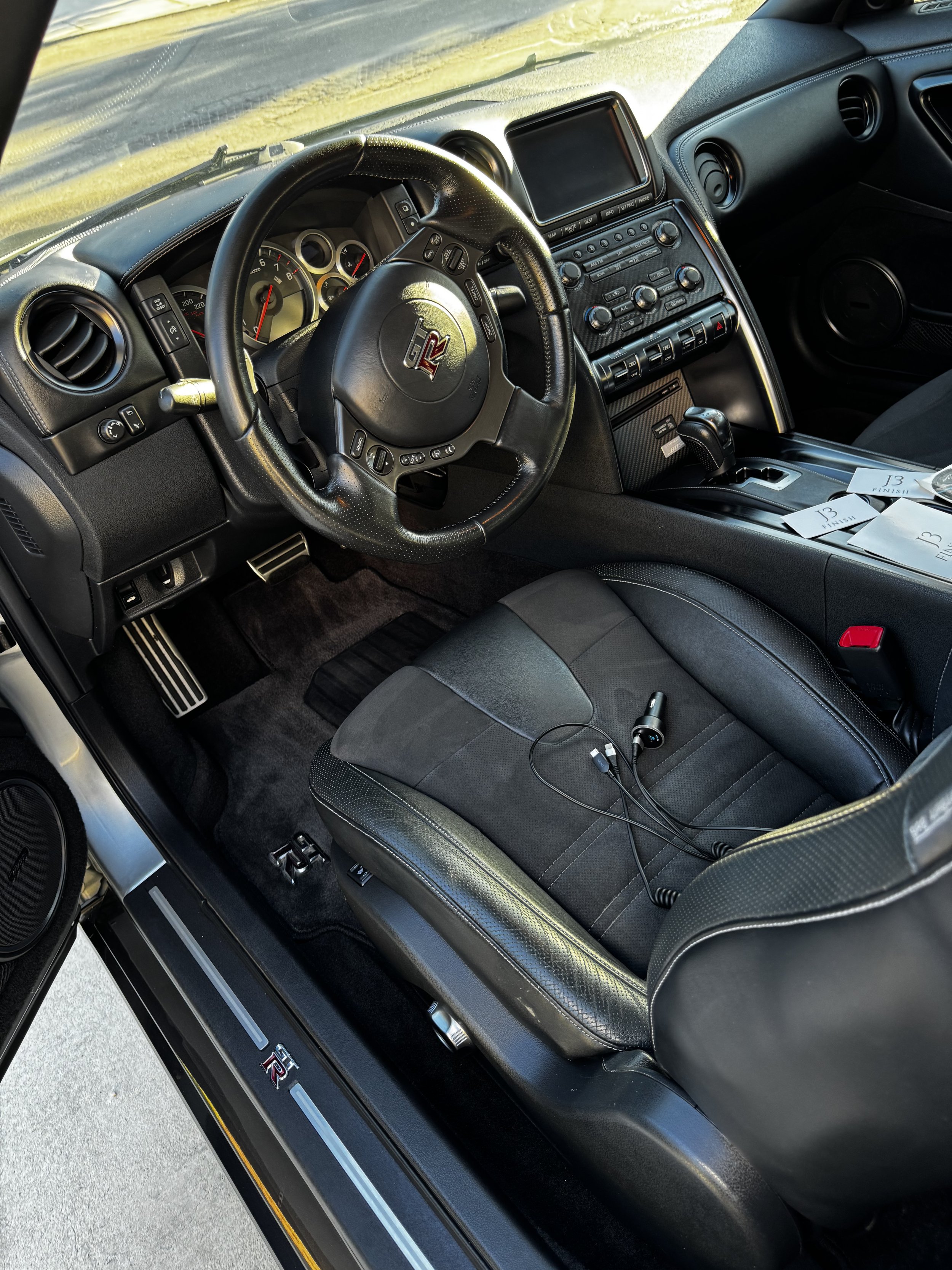 The interior of a black sports car with a steering wheel featuring a GT-R logo, digital and analog gauges, a center console with controls, and a gear shifter. There are also papers and a device connected with cords on the seat.