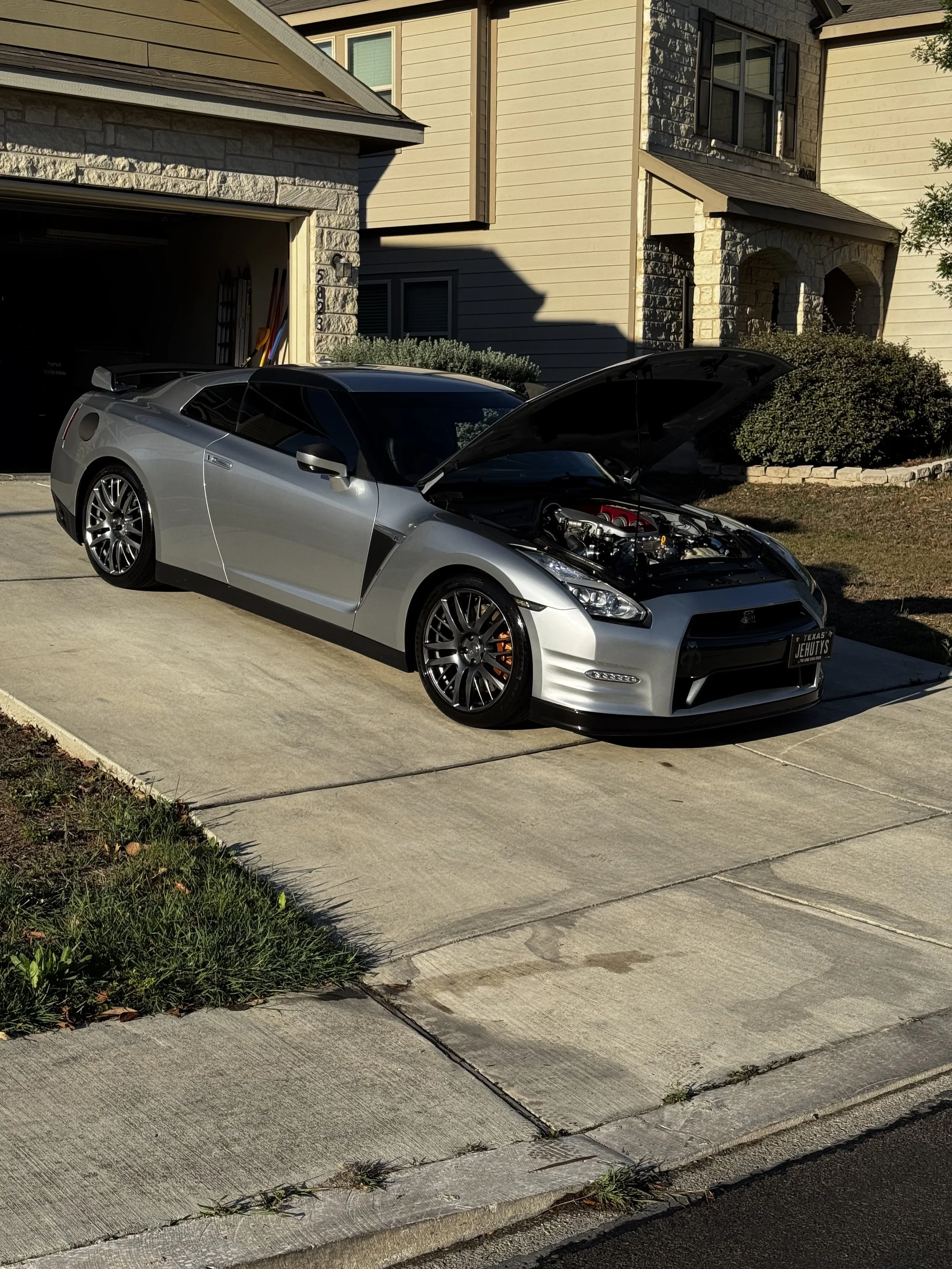 A silver sports car with its hood open, parked in front of a beige house with a garage and shrubs.