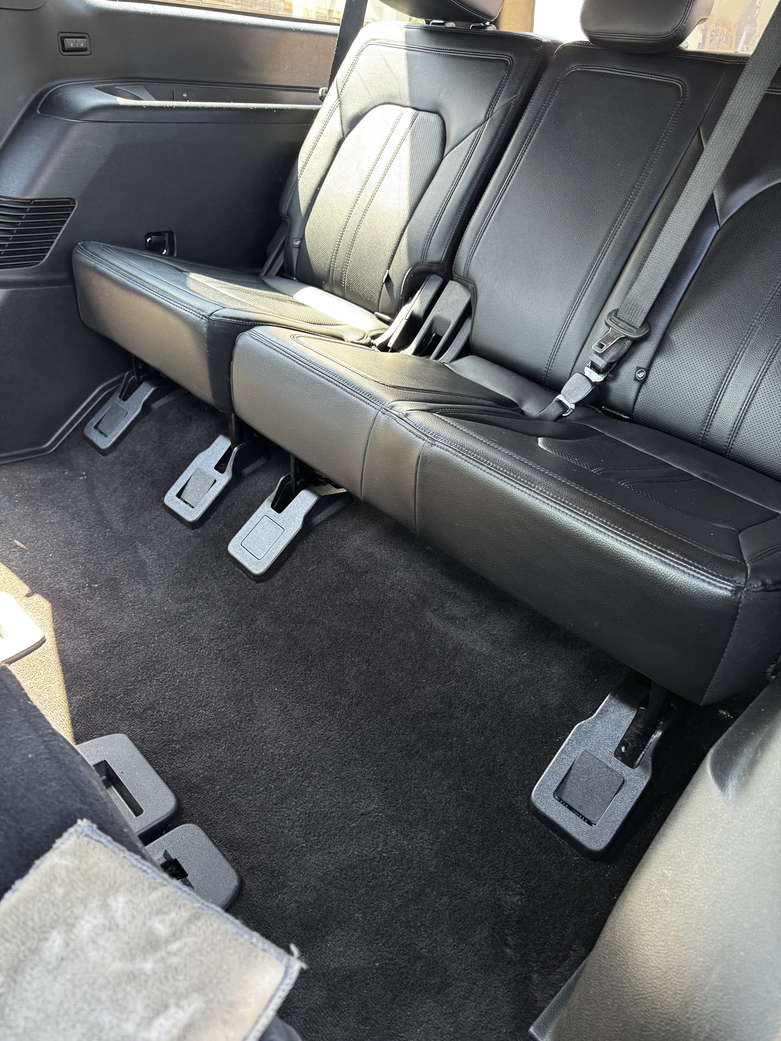 The back seats of a vehicle, showing black leather upholstery and the floor with black carpeting, with visible seatbelt hardware and pedals.