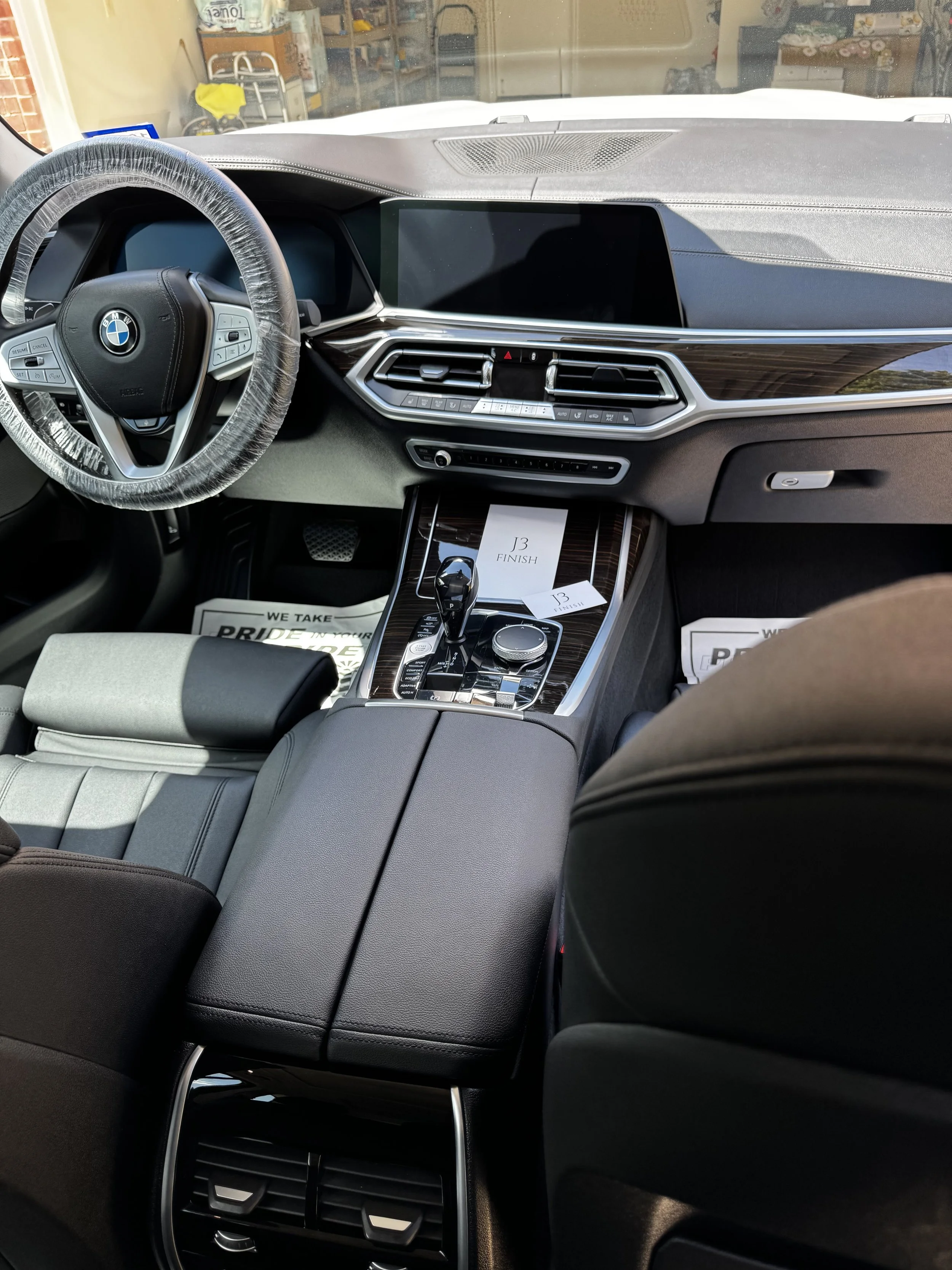 Interior of a BMW vehicle showing the steering wheel, dashboard with a large touchscreen display, center console with gear shifter and control buttons, and front passenger seat. The steering wheel is wrapped in plastic, and there are papers on the center console.