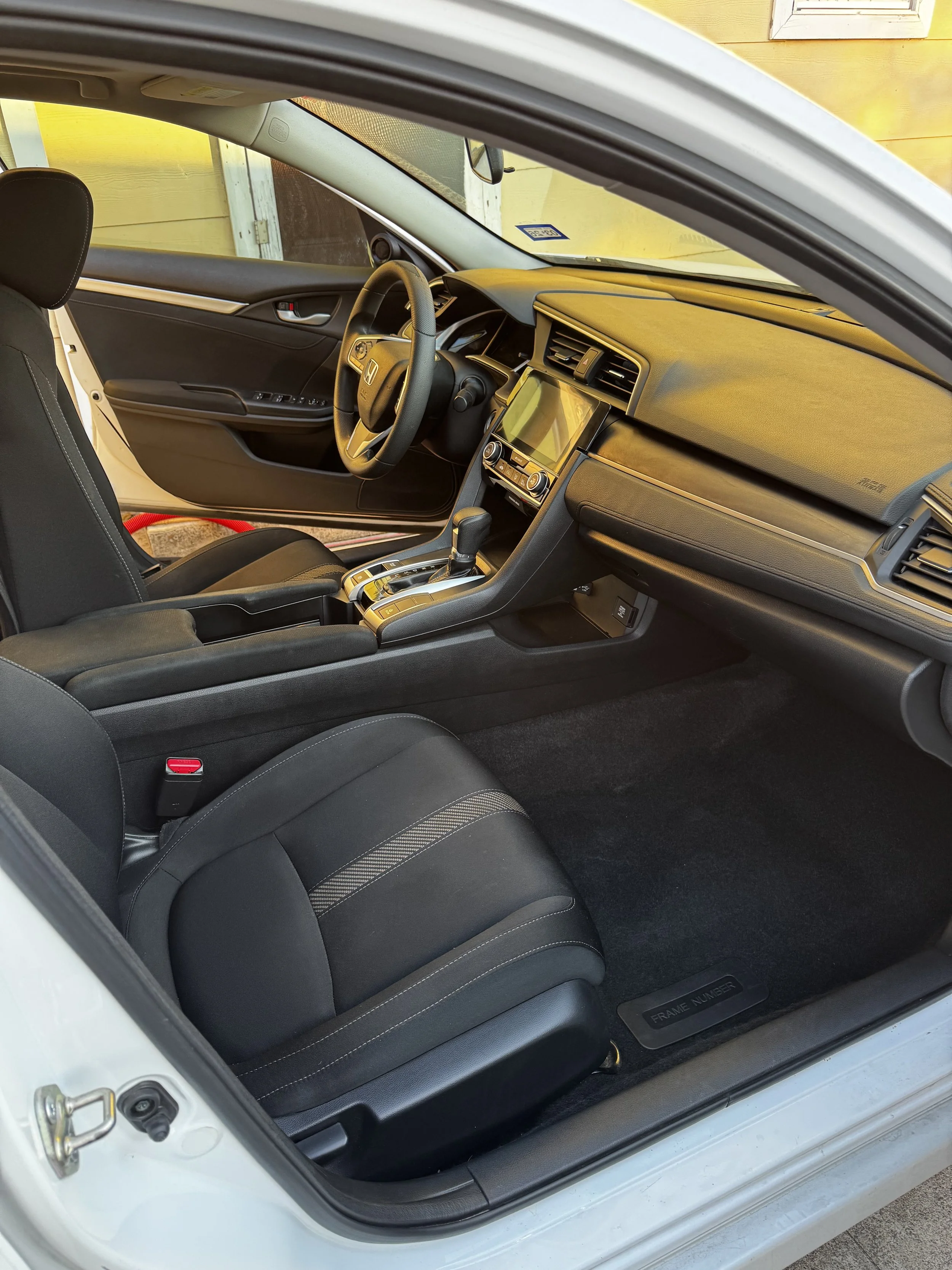 The interior of a car showing the driver's seat, steering wheel with Honda logo, dashboard, infotainment system, gear shift, and black seats with detailed stitching.