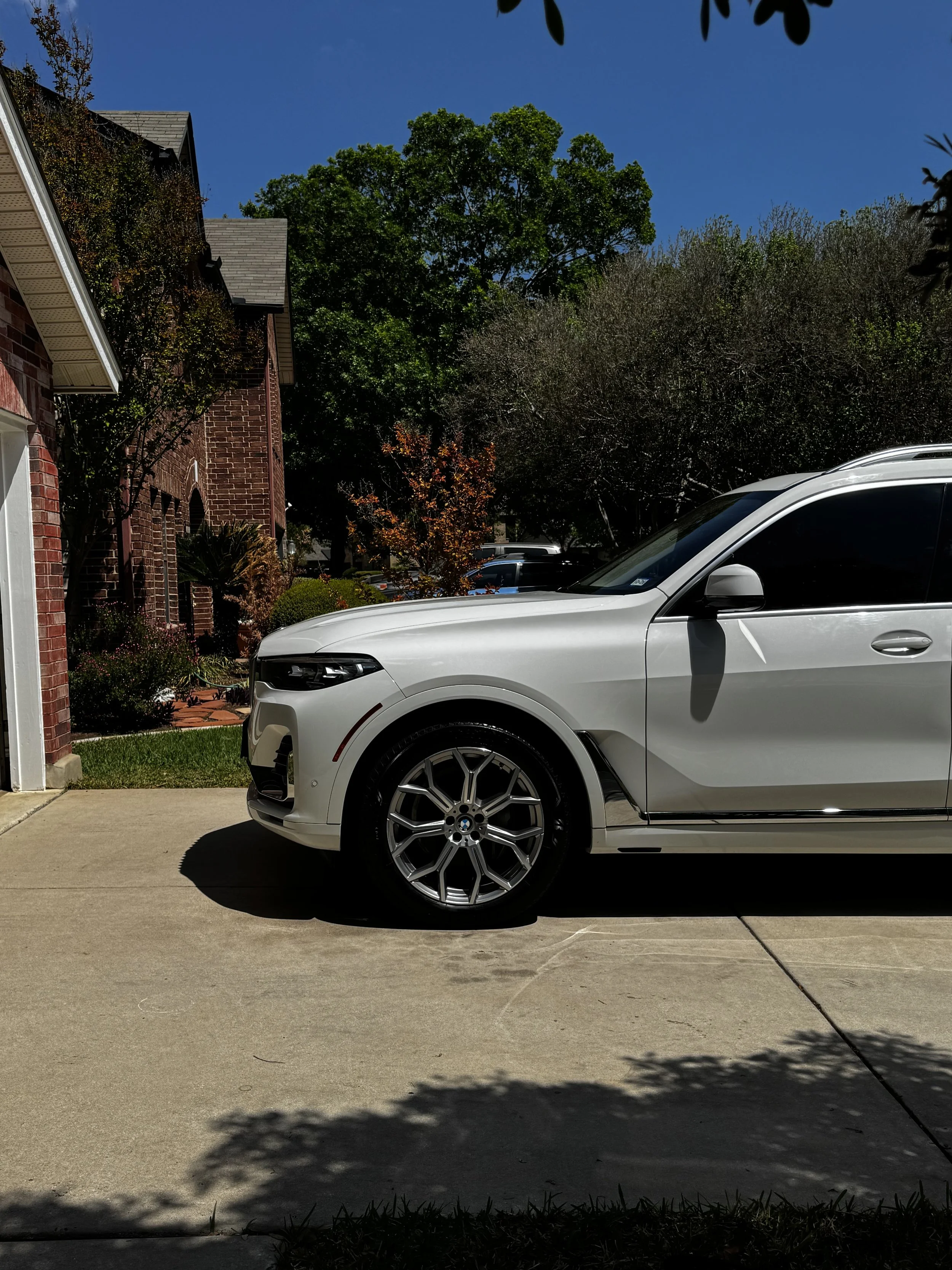 A white car parked on a driveway in front of a house with a brick exterior, surrounded by trees and plants under a blue sky.
