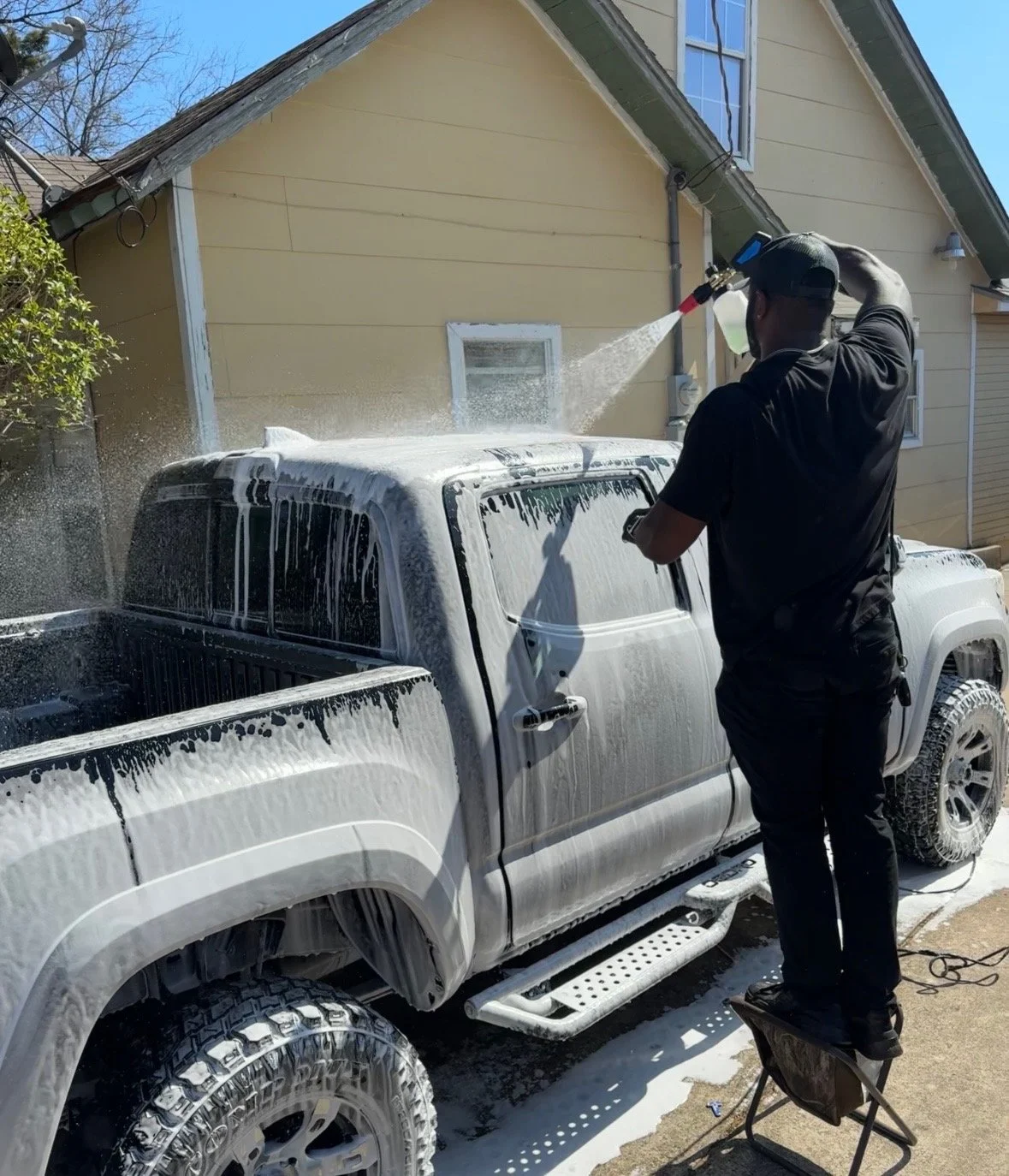 A person washing a black pickup truck outdoors with soap and water, standing on a small step stool.