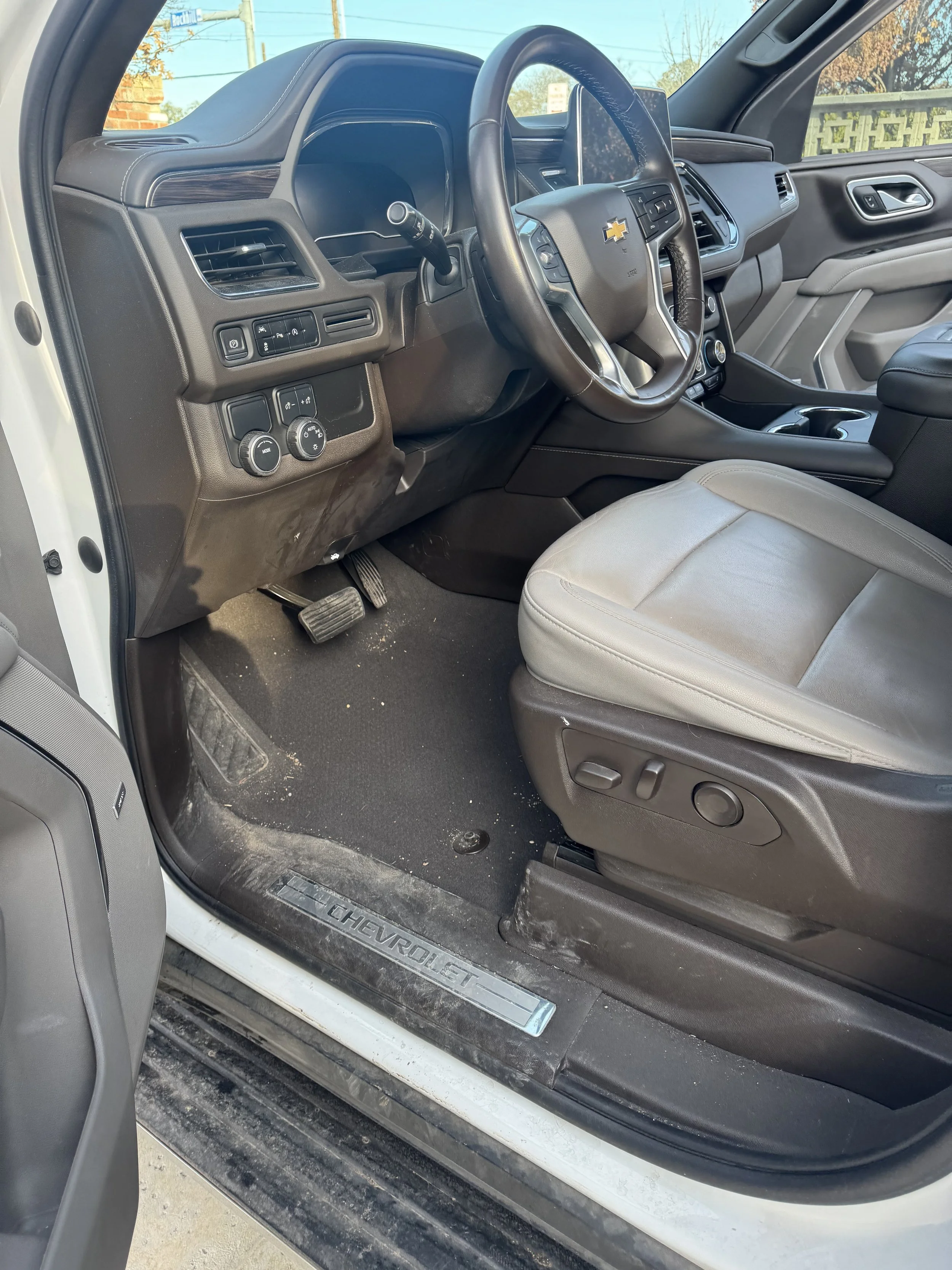 The interior front passenger side of a silver Chevrolet vehicle showing the driver's seat, steering wheel, dashboard, and door with some dirt on the floor and door sill.